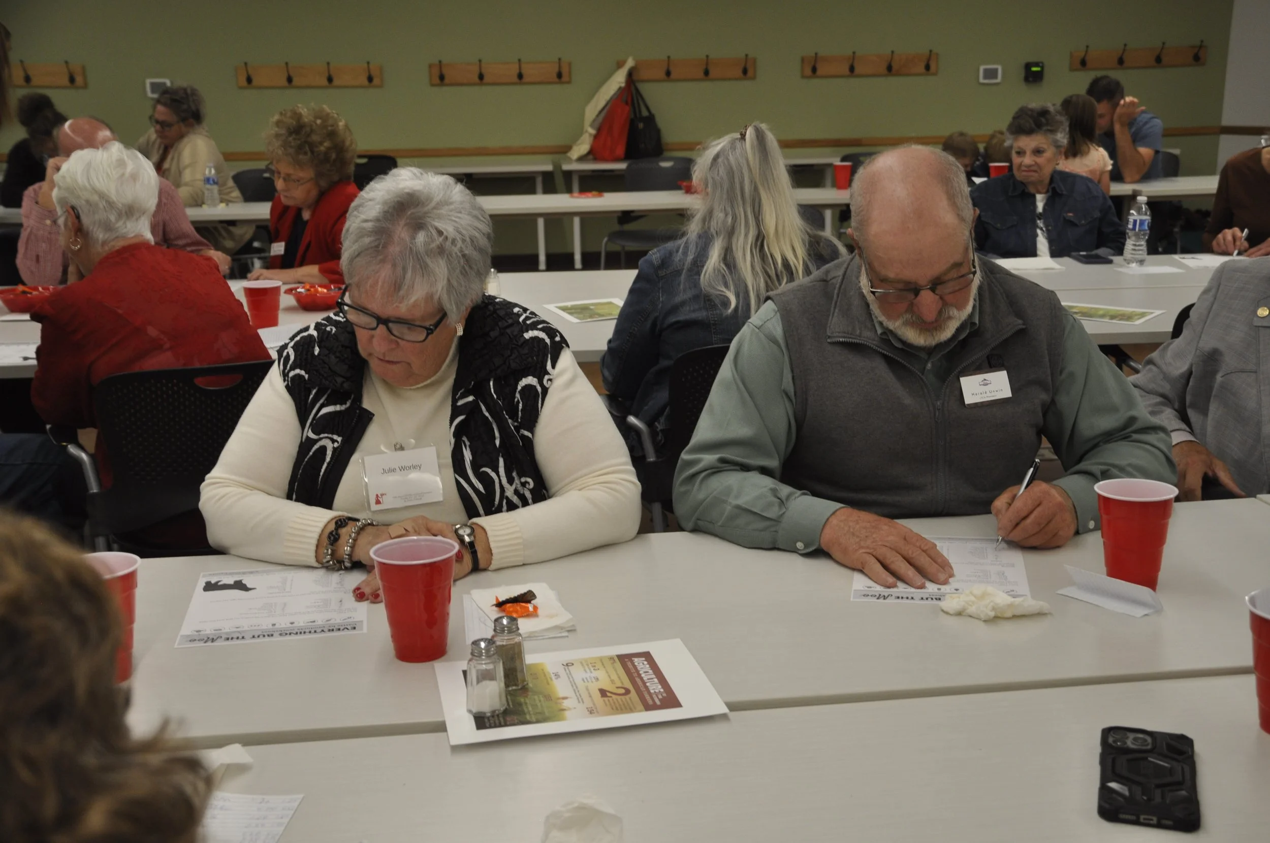 People sitting at a conference table, reading papers, with red cups, salt and pepper shakers, and pamphlets, in a room with a green wall and hooks.