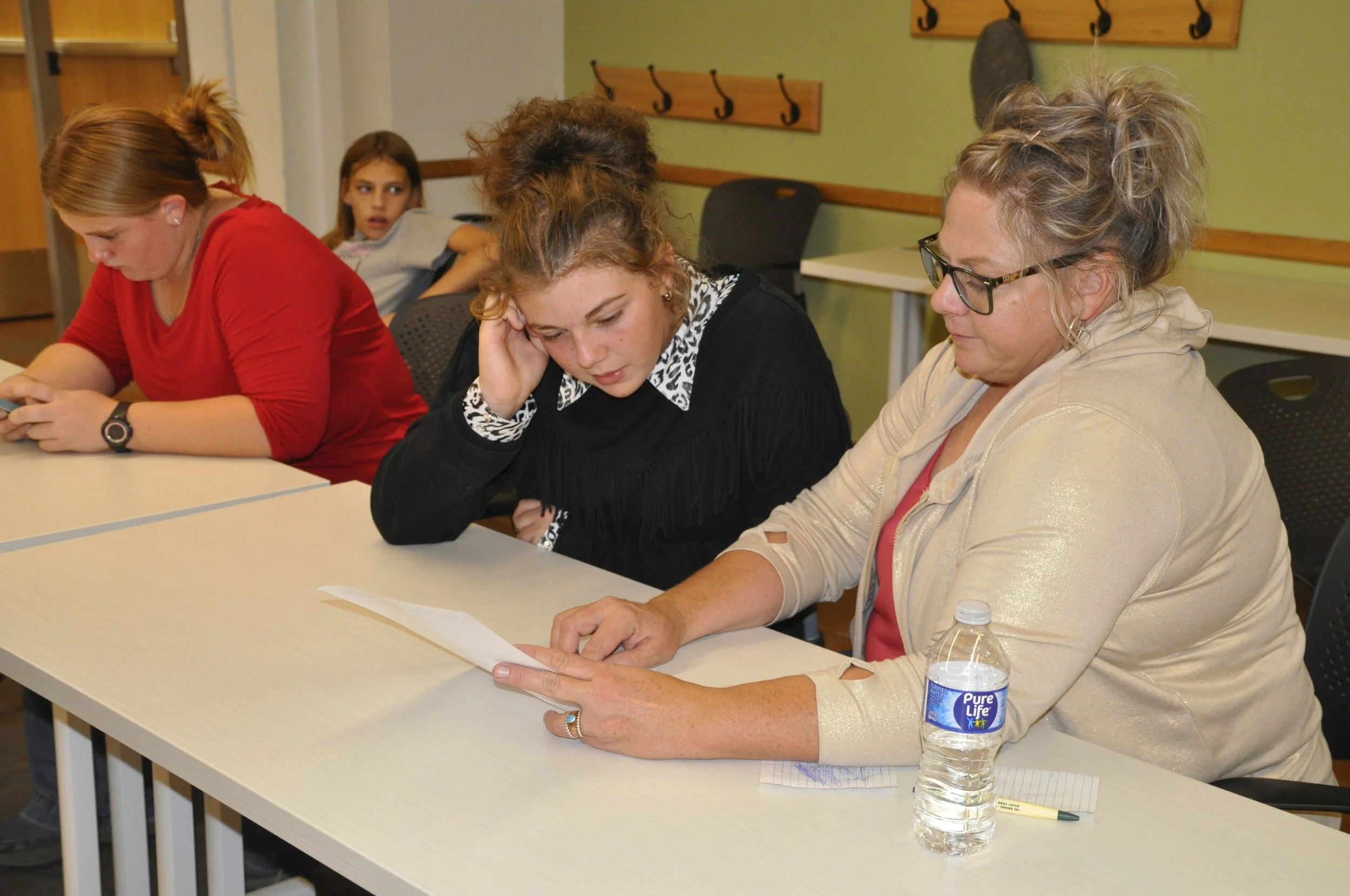 Four women sitting at a table in a classroom or meeting room. One woman is reading a paper, and the others are looking at their devices or papers. A water bottle is on the table.
