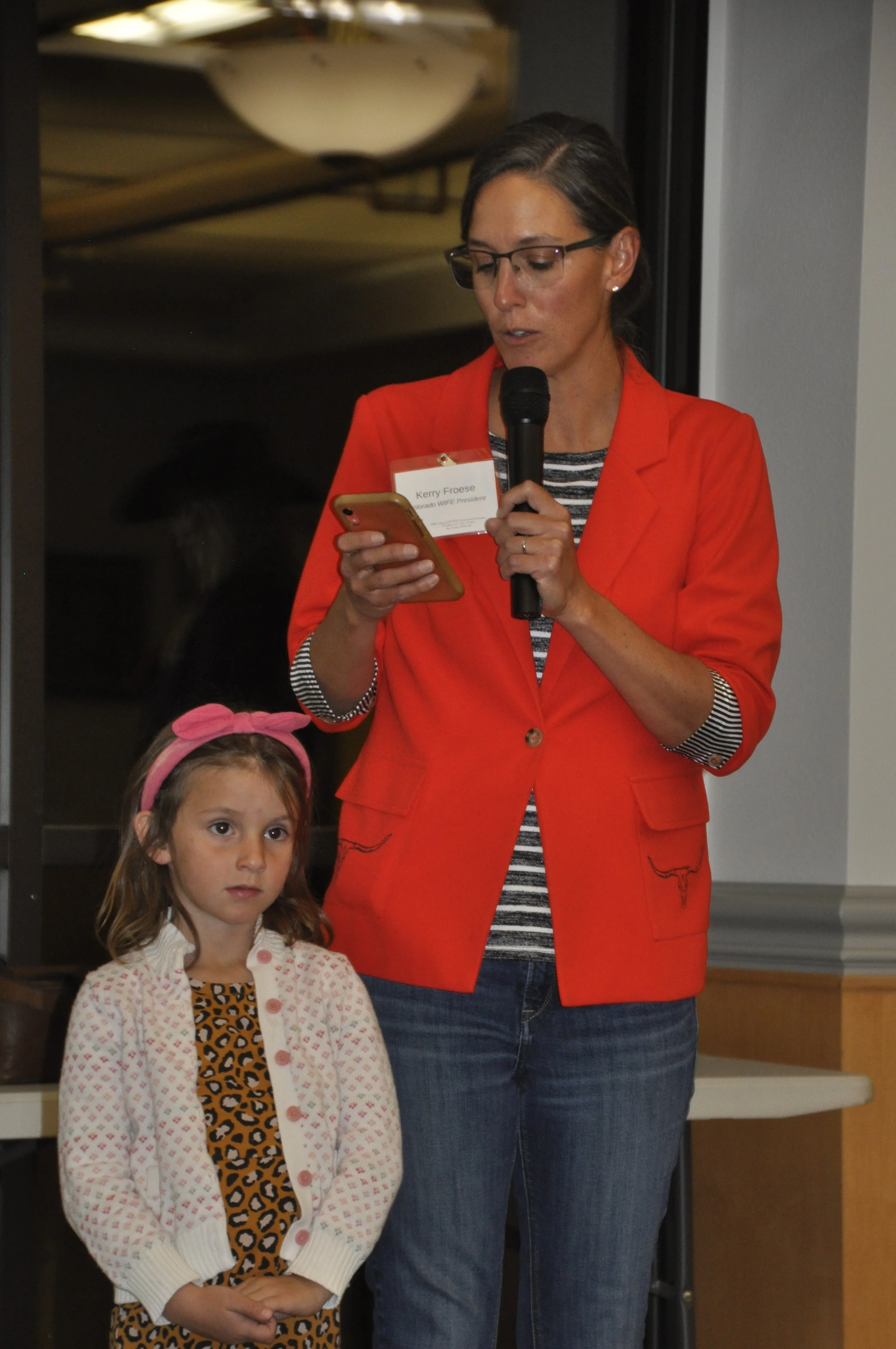 A woman wearing glasses, a red blazer, and a name tag reading "Kerry Froese, Manitoba WIFE President," is speaking into a microphone while looking at her phone. A young girl with a pink headband and a leopard print dress with a white cardigan is stan