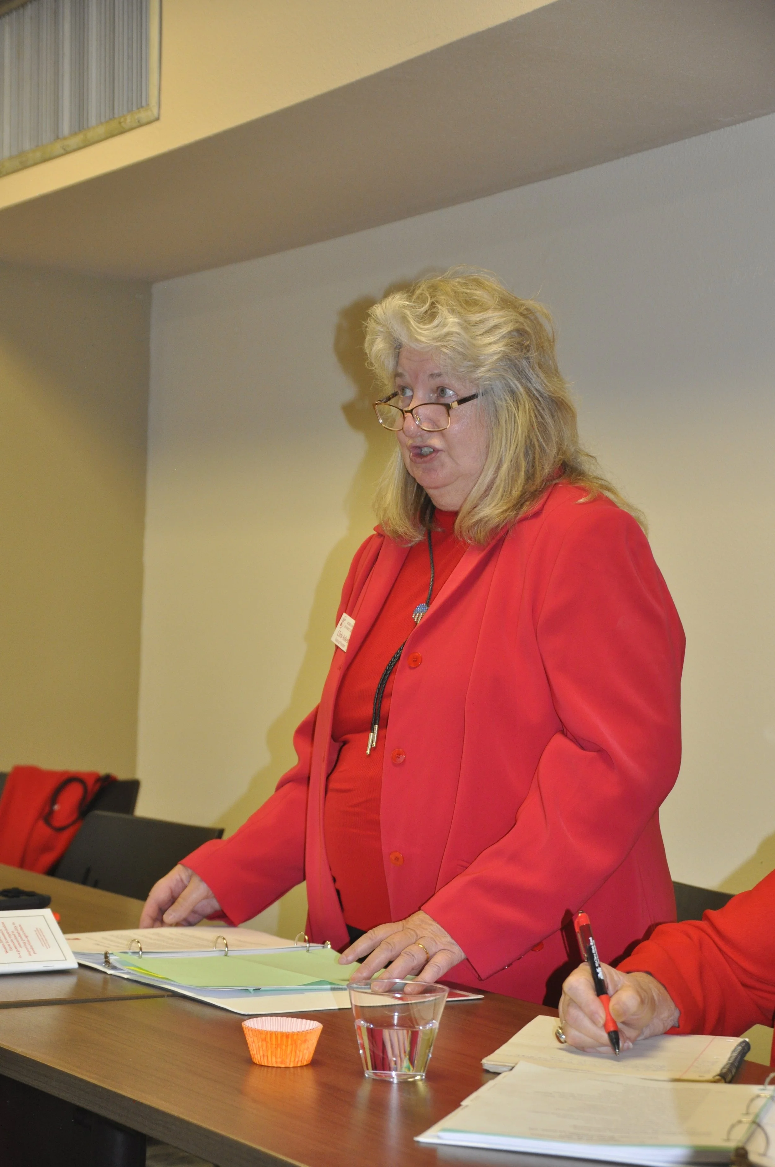 A woman with blonde curly hair, glasses, and a red blazer standing at a brown table with papers, a glass of water, and a person sitting to her right. The woman appears to be speaking, and the setting looks like a meeting or conference room.