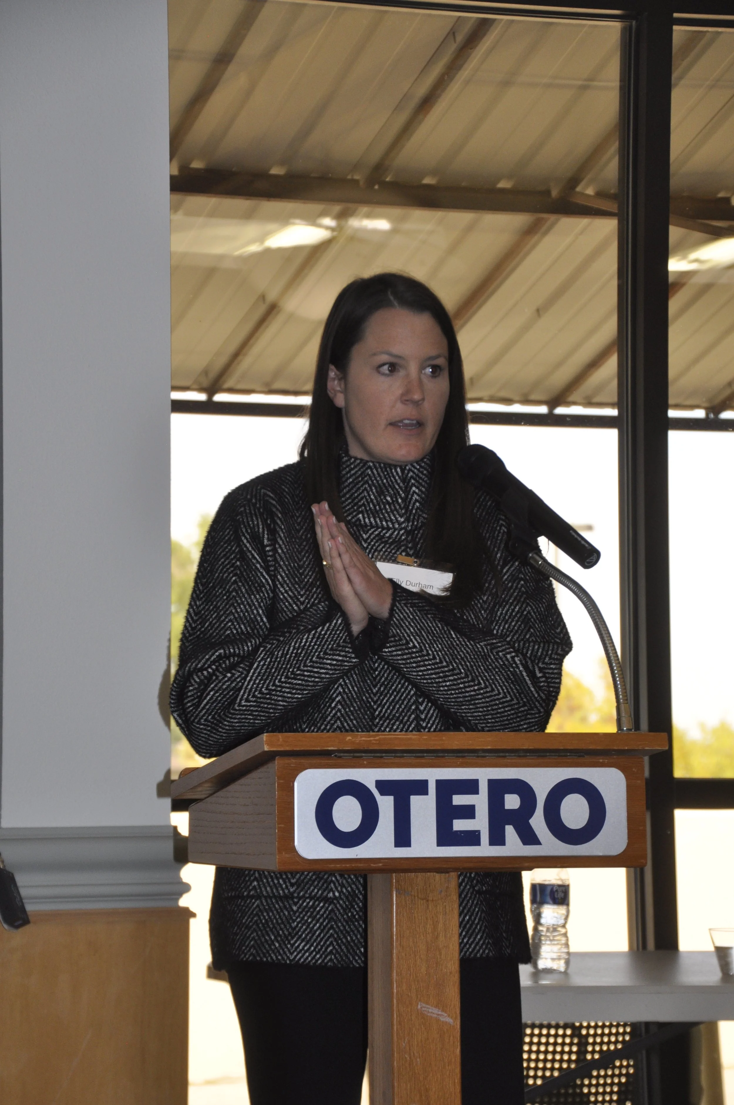 A woman with long dark hair speaking at a podium with a microphone. She is wearing a black and white patterned jacket and has her hands clasped near her chest. There is a sign on the podium that says 'OTERO'. In the background, there is a window show