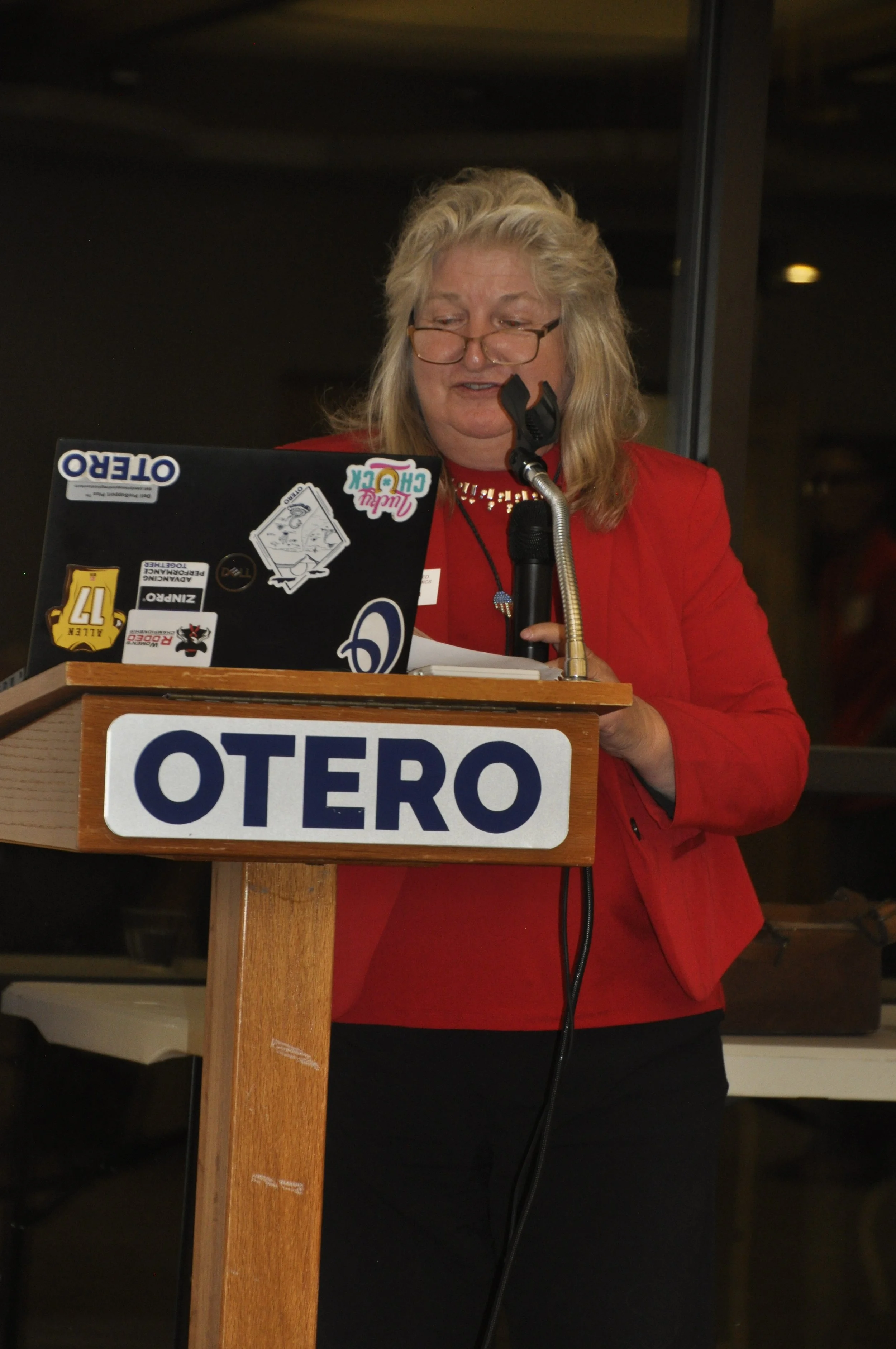 A woman with blonde hair wearing glasses and a red blazer standing at a wooden podium with stickers on a laptop and a sign that reads 'OTERO', speaking into a microphone.