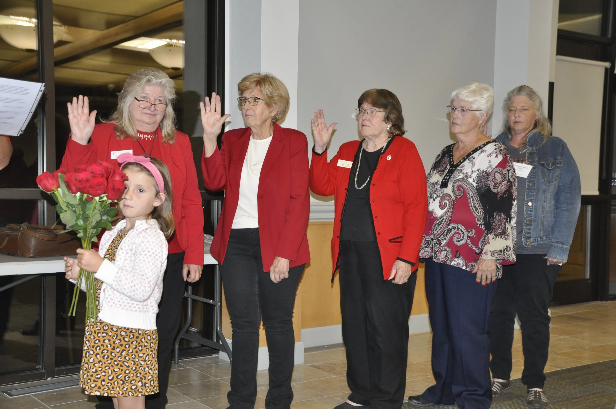 A group of six women and one young girl are standing indoors, with five women raising their right hands as if taking an oath. The young girl in front is holding a bouquet of red roses and looking to the side. The women are wearing casual to semi-form