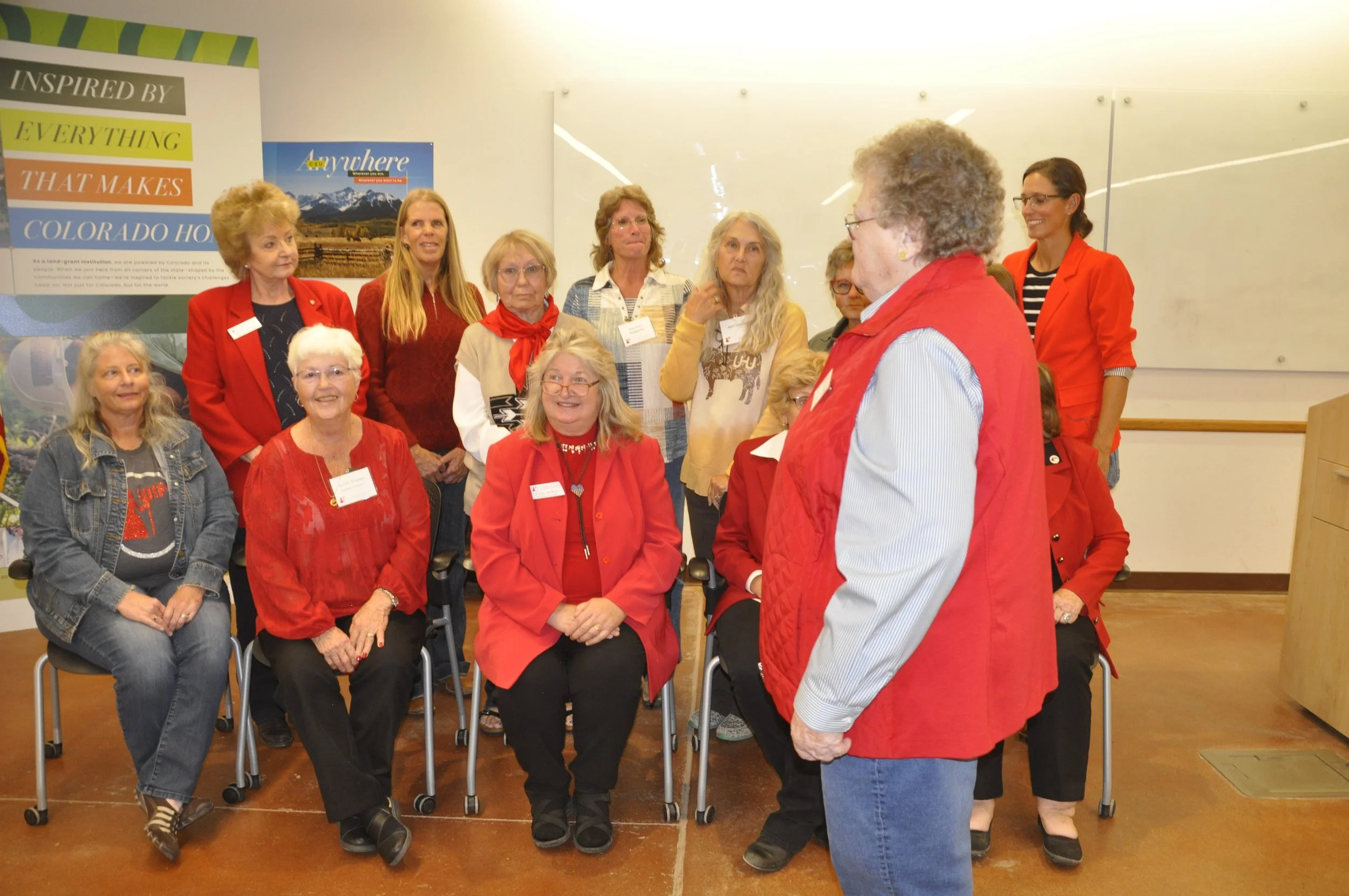 Group of women gathered in a room, some seated and some standing, engaged in conversation. Many are wearing red or red clothing. The background features a whiteboard and posters, including one related to Colorado.