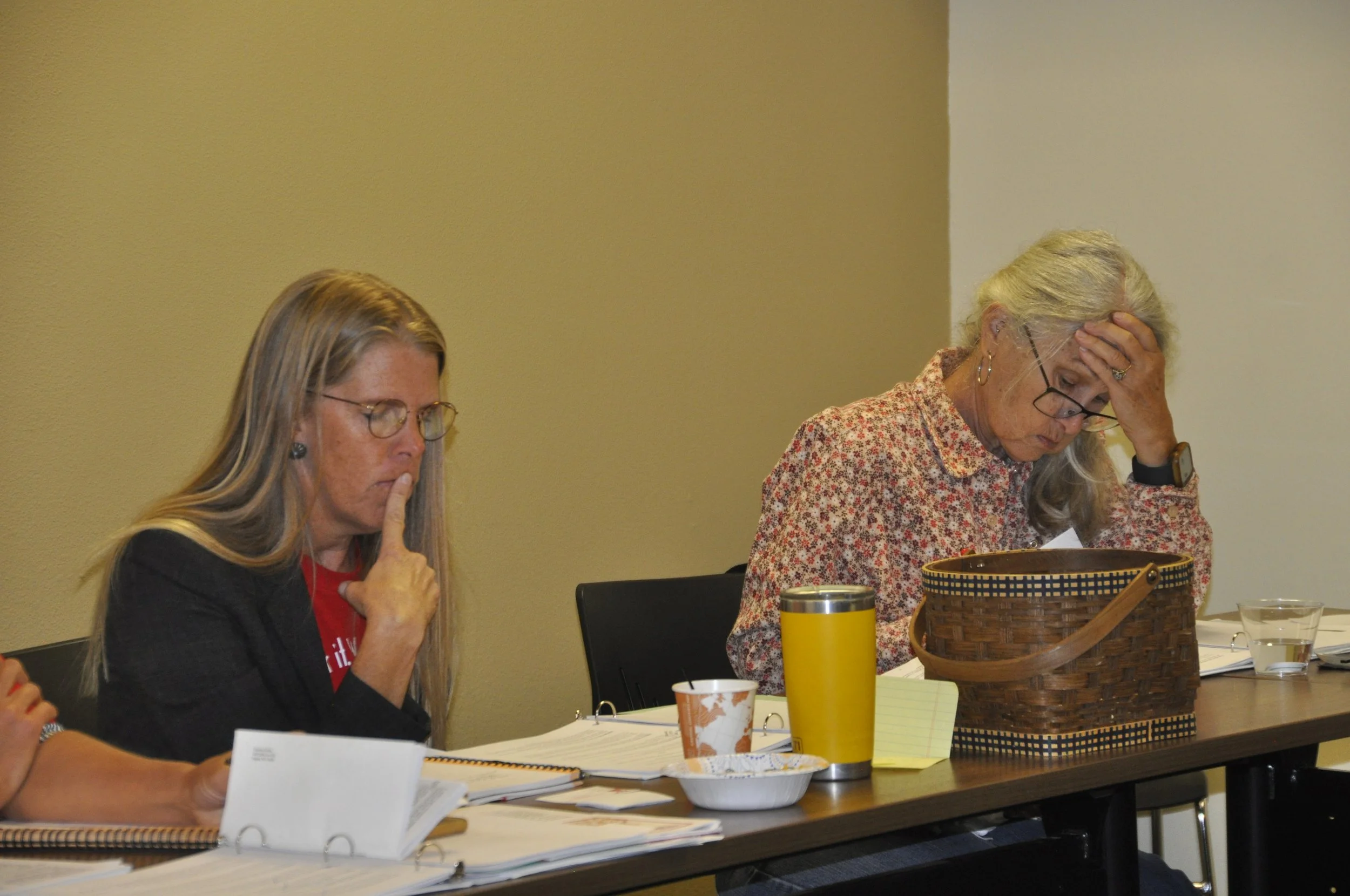 Two women with glasses sitting at a table, reading papers, with a basket, cups, and a yellow thermos on the table; one woman with blonde hair in a black jacket, the other with gray hair and a patterned shirt, appears to be distressed.