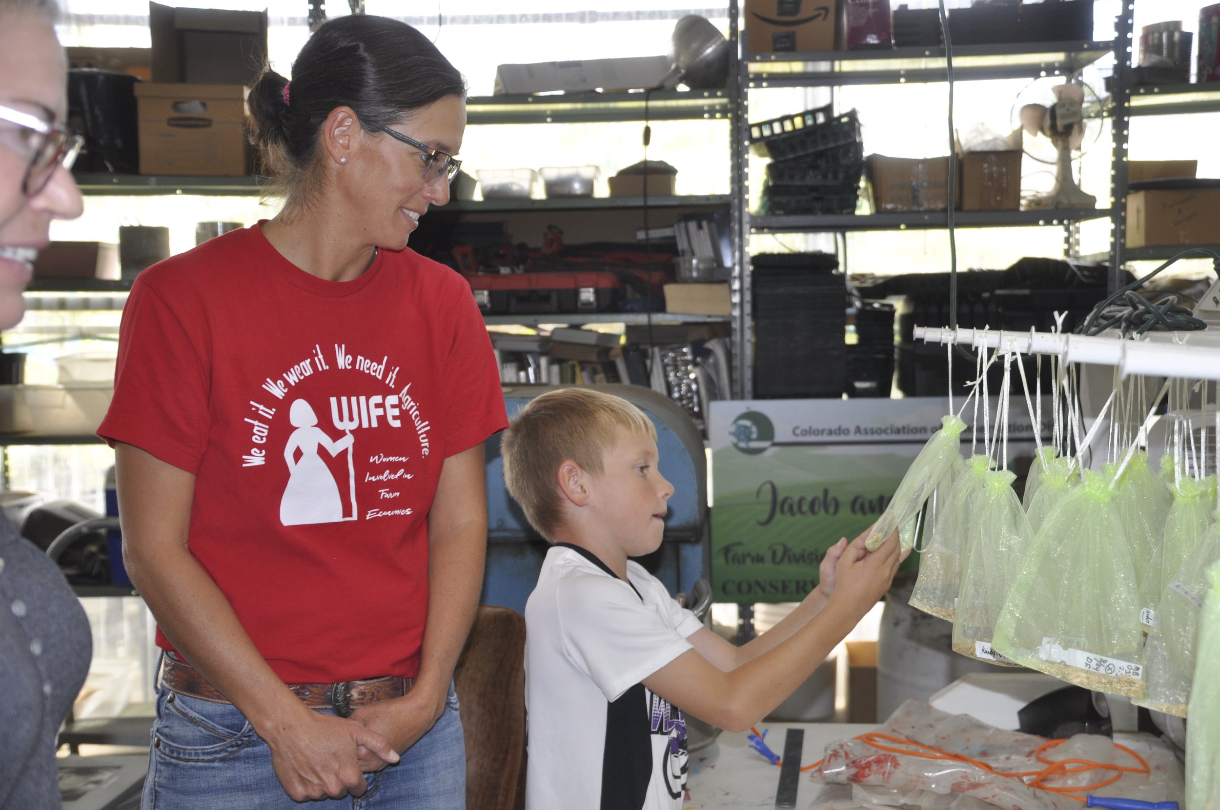 A woman wearing glasses and a red t-shirt with white text and a logo, standing next to a young boy with a white shirt, as they look at items in clear plastic bags at a table in an indoor setting with shelves and boxes in the background.