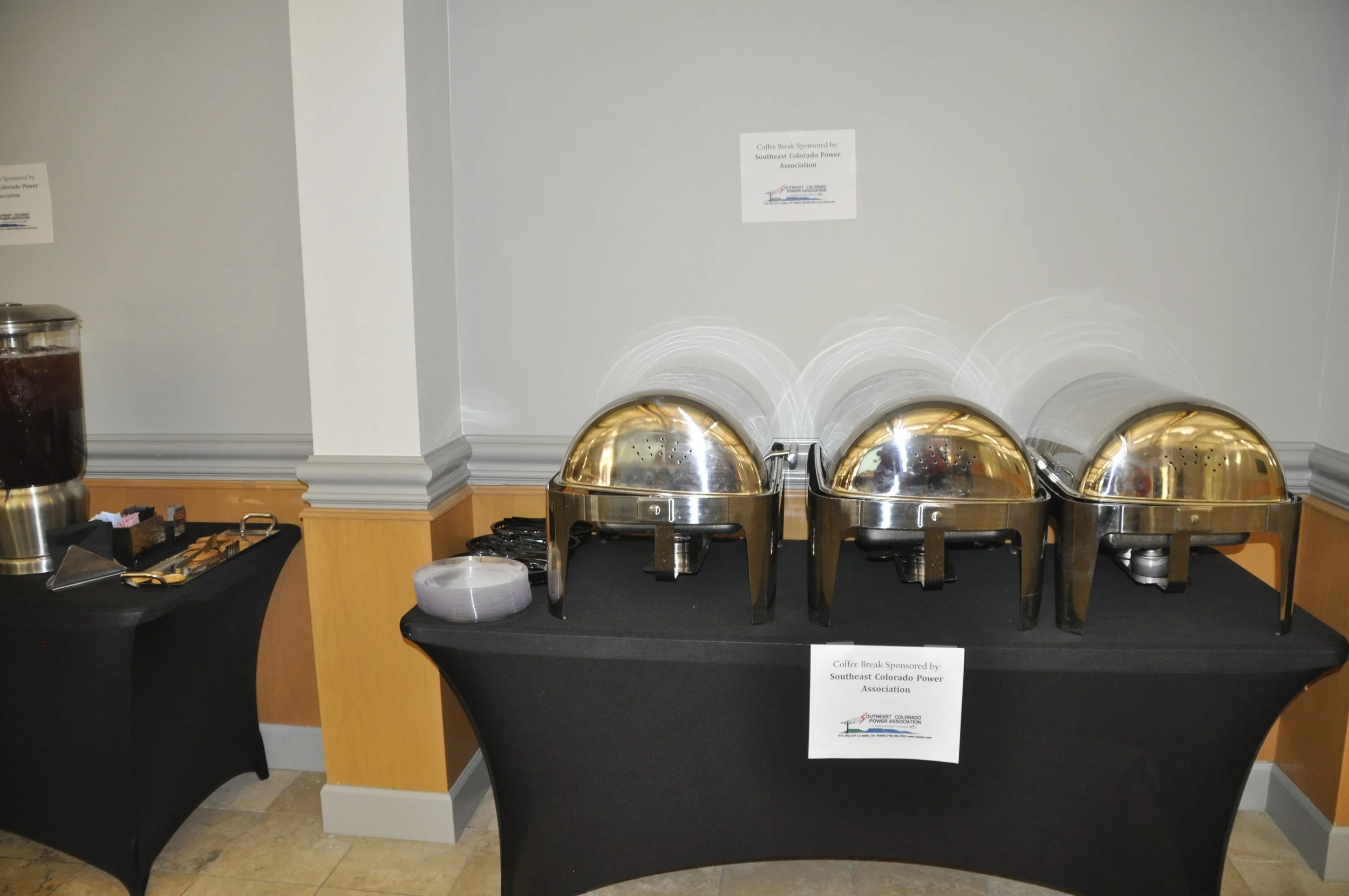 Buffet table with breakfast chafing dishes, a coffee pot, napkins, and condiments, set against a neutral wall with small signage indicating sponsorship by the Southeast Colorado Power Association.