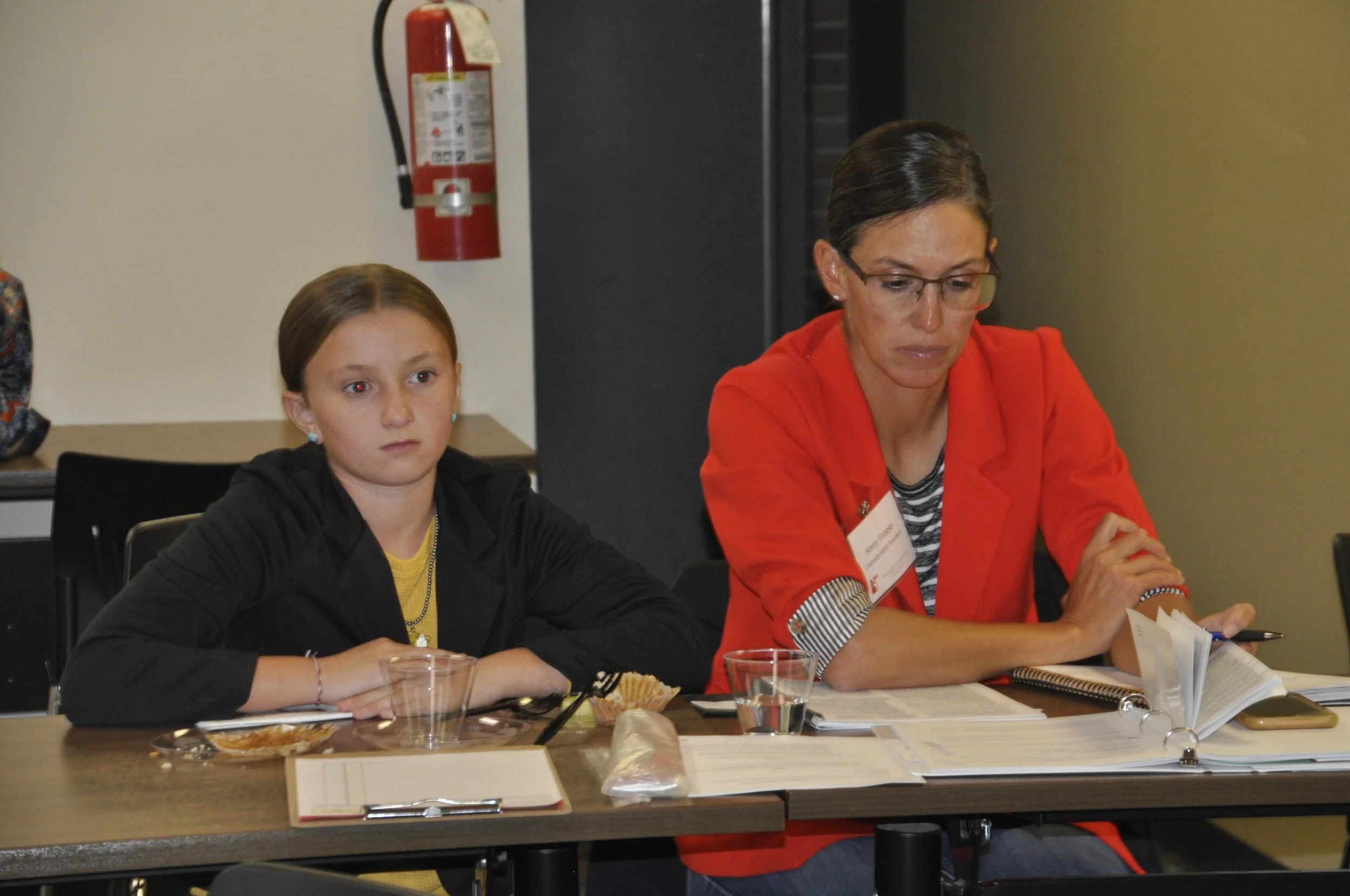 A woman and a young girl sitting at a table during a meeting or discussion, with papers, notebooks, and food in front of them.