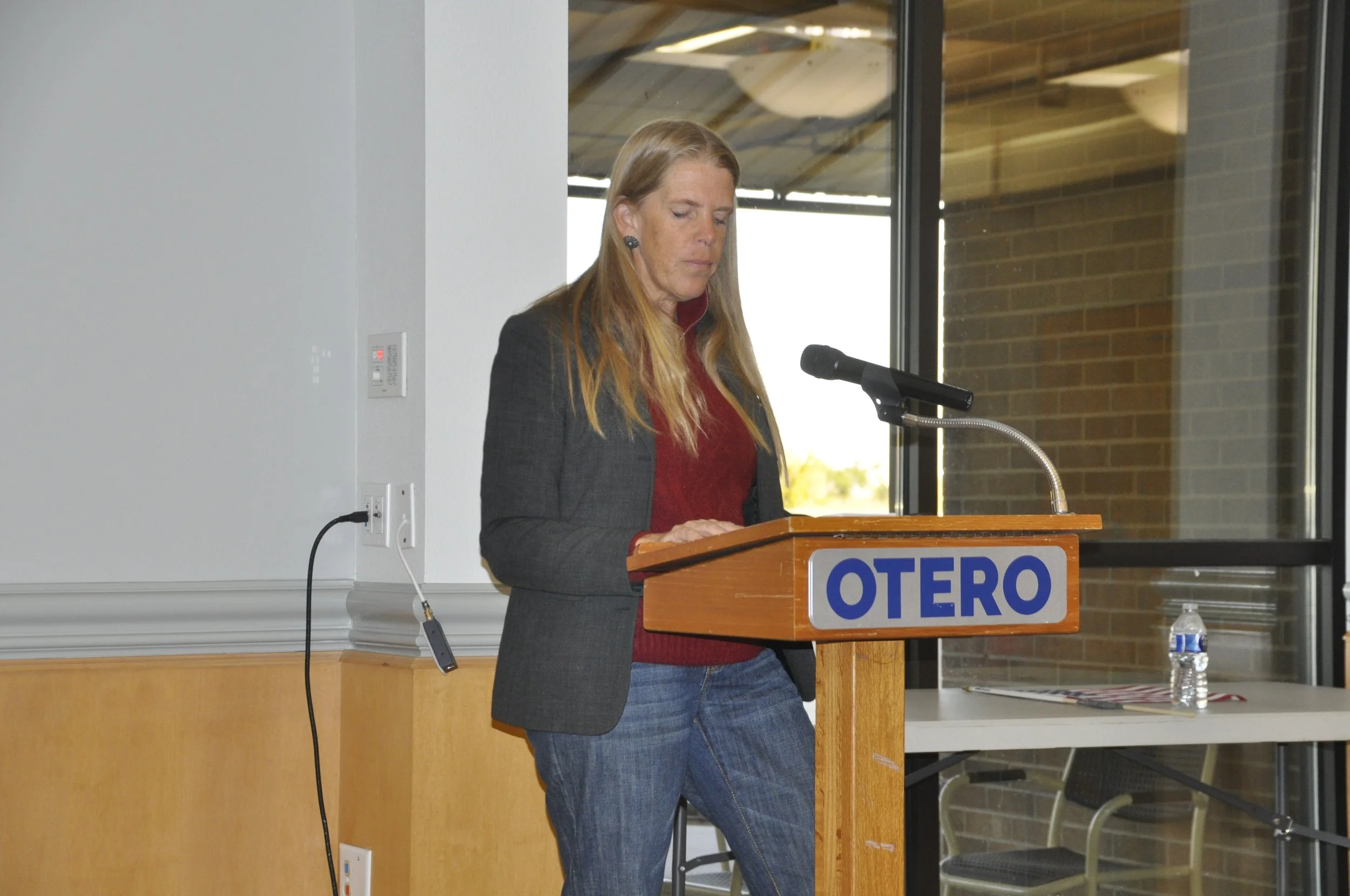 A woman with long blonde hair standing at a podium labeled "OTERO", reading or speaking into a microphone in a room with large windows and brick walls.