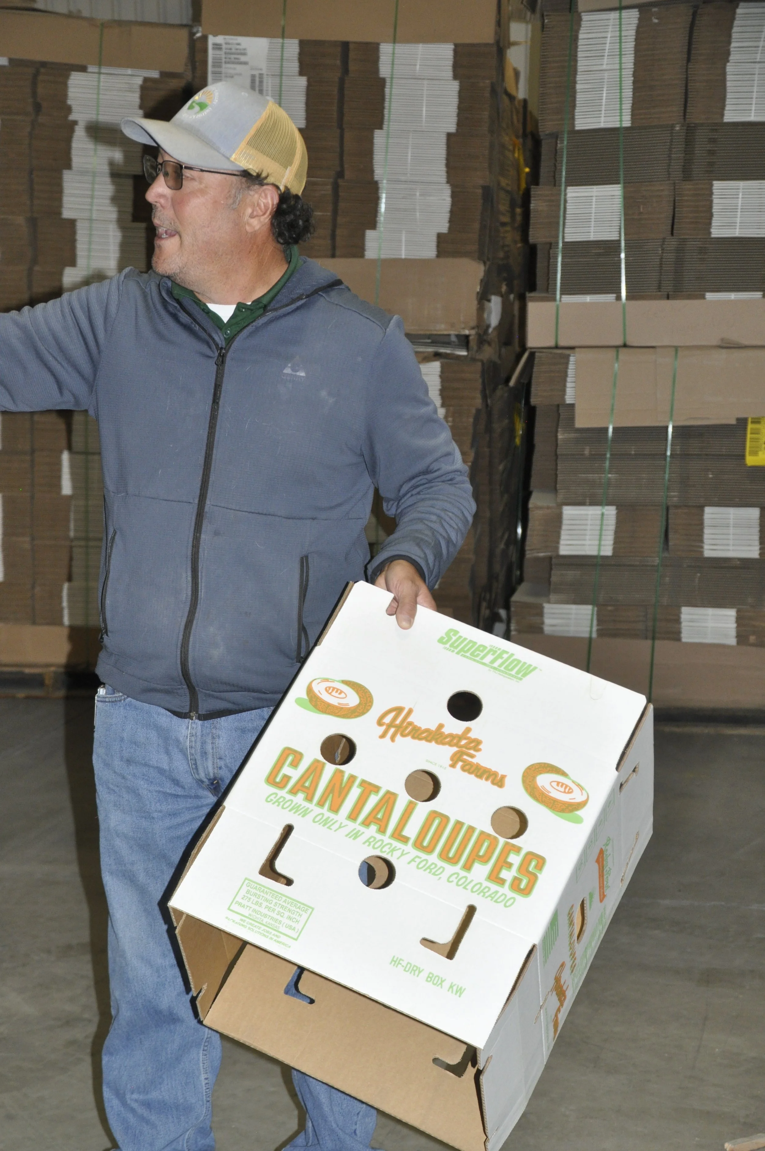 A man wearing glasses, a gray cap, and a gray jacket is carrying a large box labeled 'Cantaloupe' in a warehouse filled with stacks of boxes.