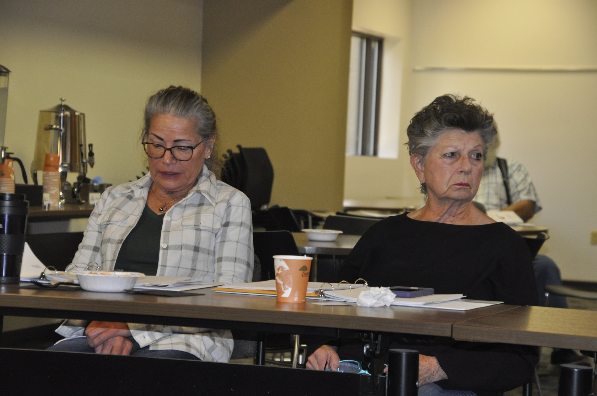 Two older women sit at a table in a room with beige walls. The woman on the left has gray hair, glasses, and is wearing a plaid shirt. The woman on the right has short, curly gray hair and is wearing a black top. The table has papers, a binder, a dis