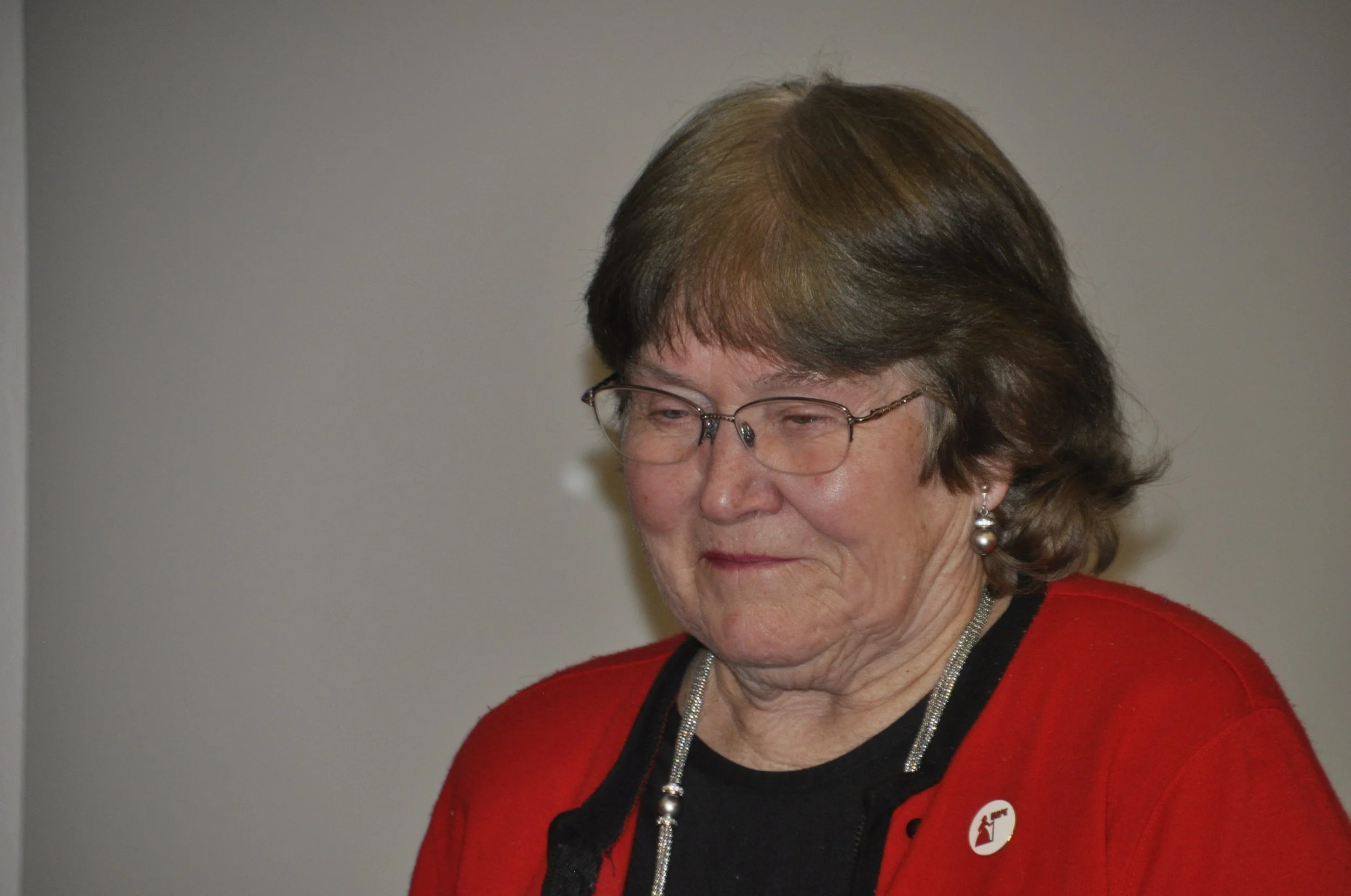 A woman with glasses and short brown hair wearing a red cardigan with a black shirt and silver jewelry, looking down with a neutral expression against a plain background.