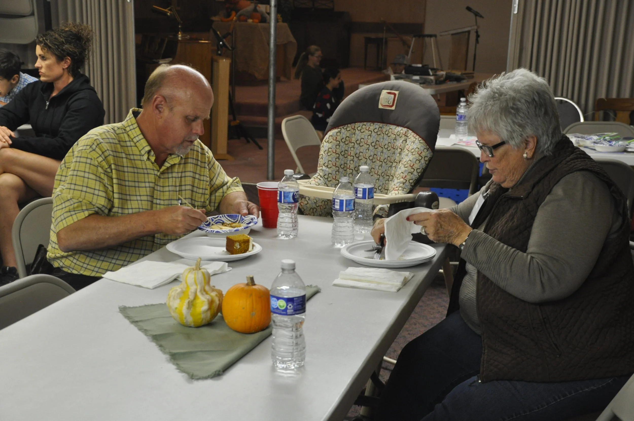Three elderly people sitting at a table with fall decorations, eating food, with water bottles, cups, and a slice of cake on a plate in front of them. Two women and one man, with pumpkins and gourds on the table.