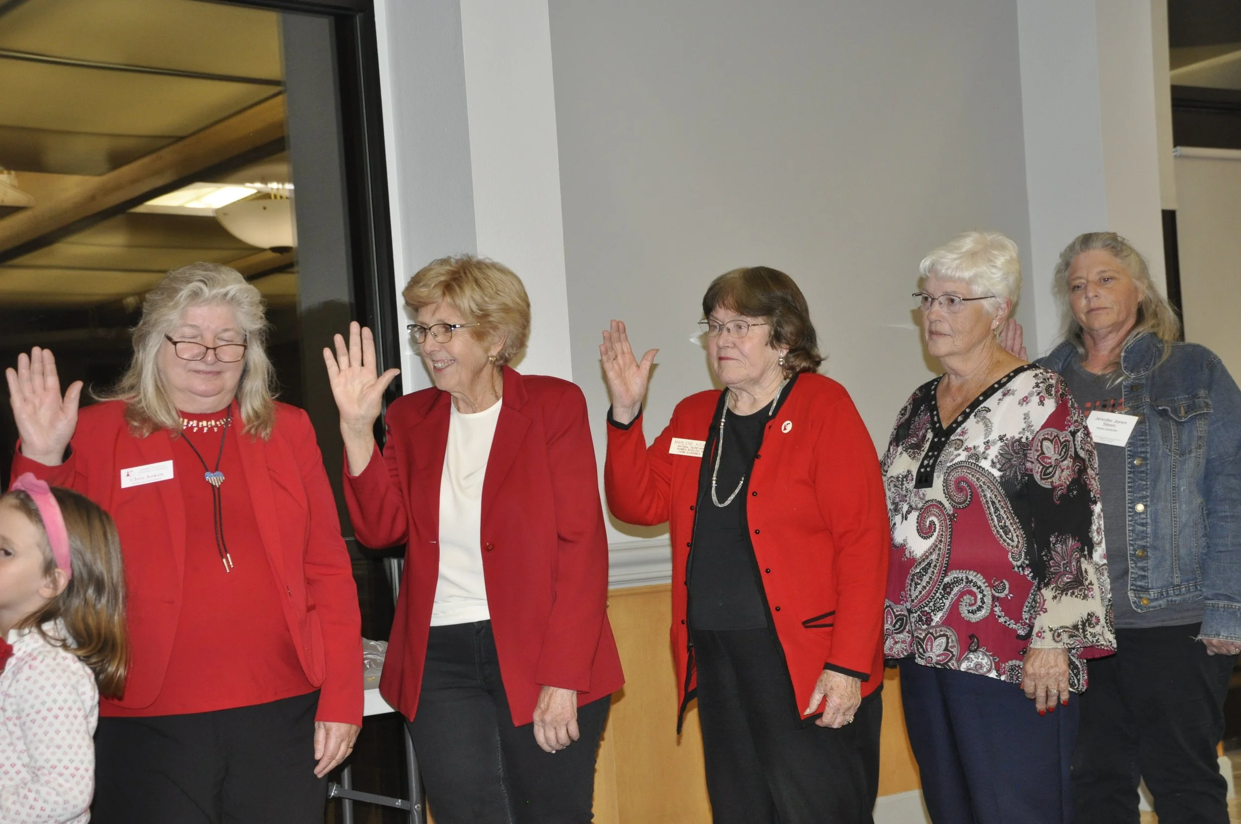 A group of older women standing indoors, some wearing red jackets or shirts, participating in an event or gathering, with a young girl in the foreground wearing a white and pink polka-dot shirt.
