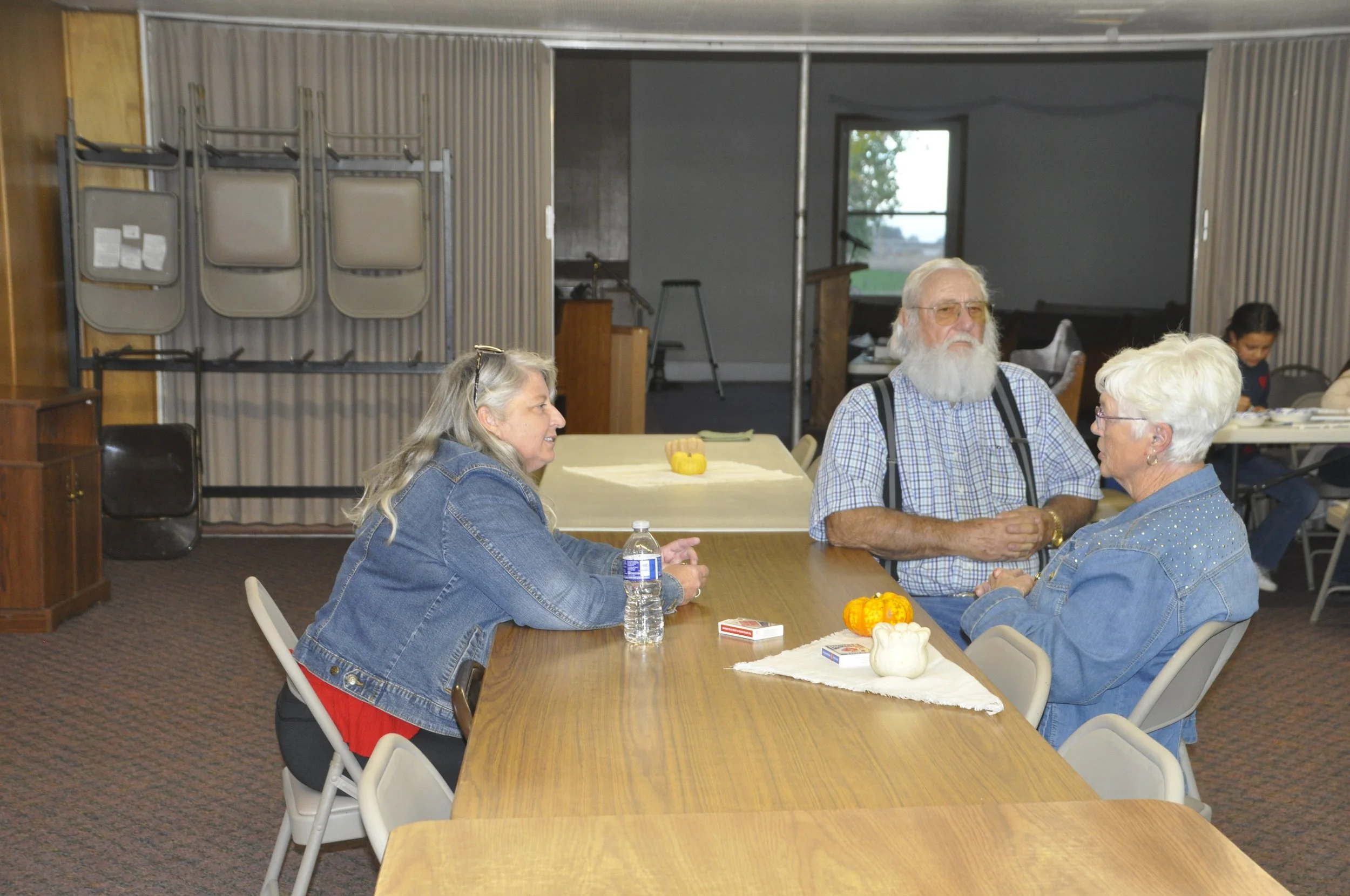 Four seniors sitting at a long table in a room, engaging in conversation. The table has small pumpkins and a water bottle. The room has a background with a window, chairs, and stacked folding tables.