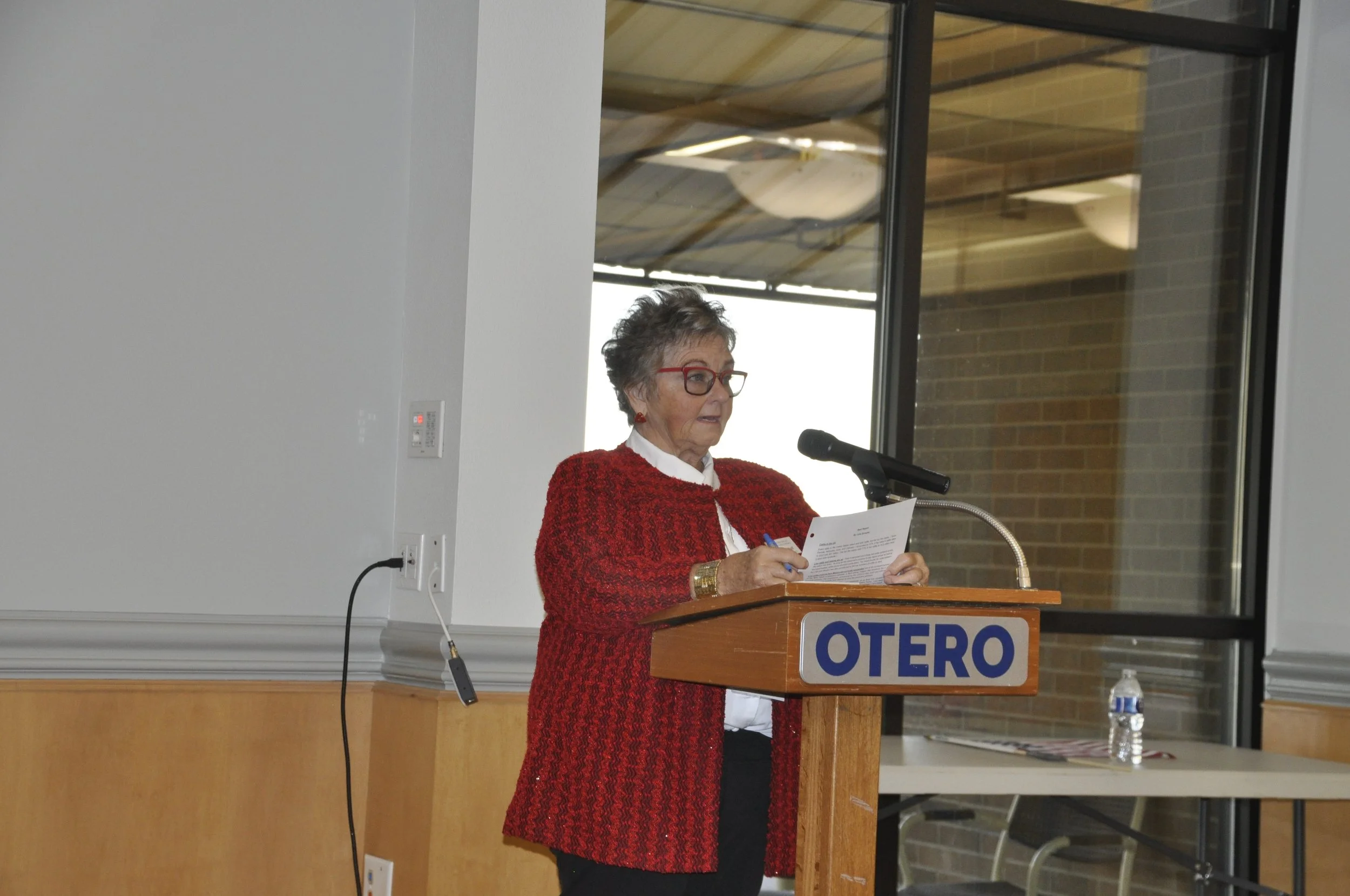 An elderly woman with glasses, wearing a red blazer, stands behind a wooden podium reading from a paper. The podium has a sign that reads 'OTERO'. There is a microphone near her mouth and a water bottle on the table to the right.