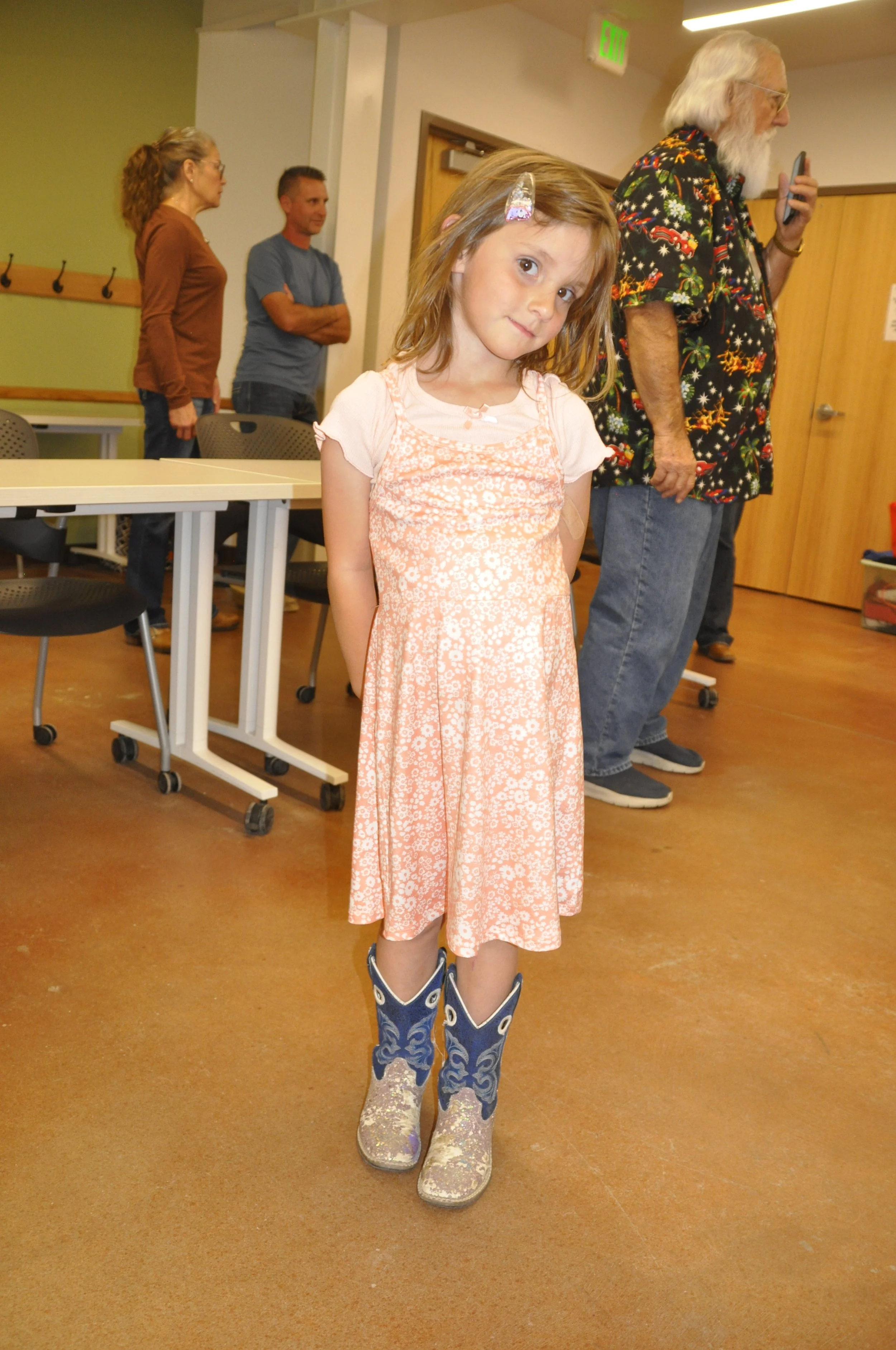 A young girl in a peach-colored floral dress standing in a room with brown floors and beige walls. She is wearing blue cowboy boots with glitter and is looking at the camera with a slight smile.