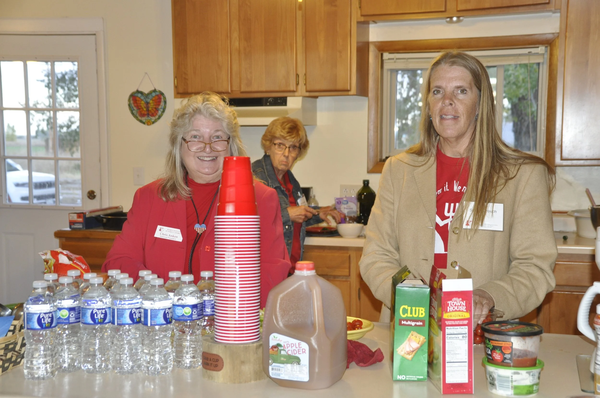 Three women standing behind a table filled with bottled water, juice, and snacks in a kitchen with wooden cabinets and a window. One woman wears glasses and a red shirt, another has red hair and glasses, and the third woman has long, straight hair an