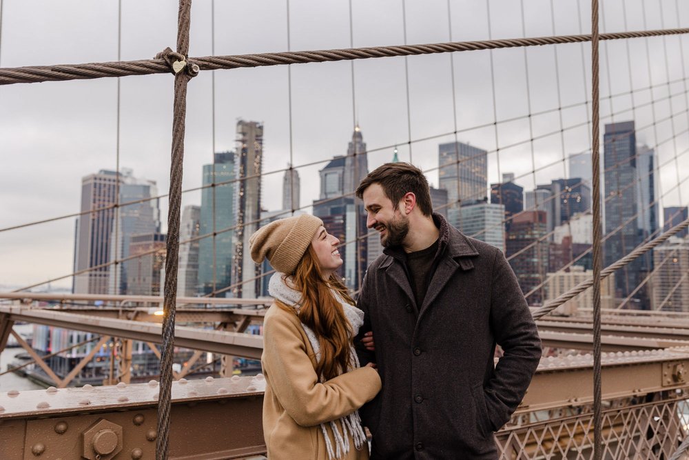 An engaged couple  smiling at each other, with the New York City skyline in the background