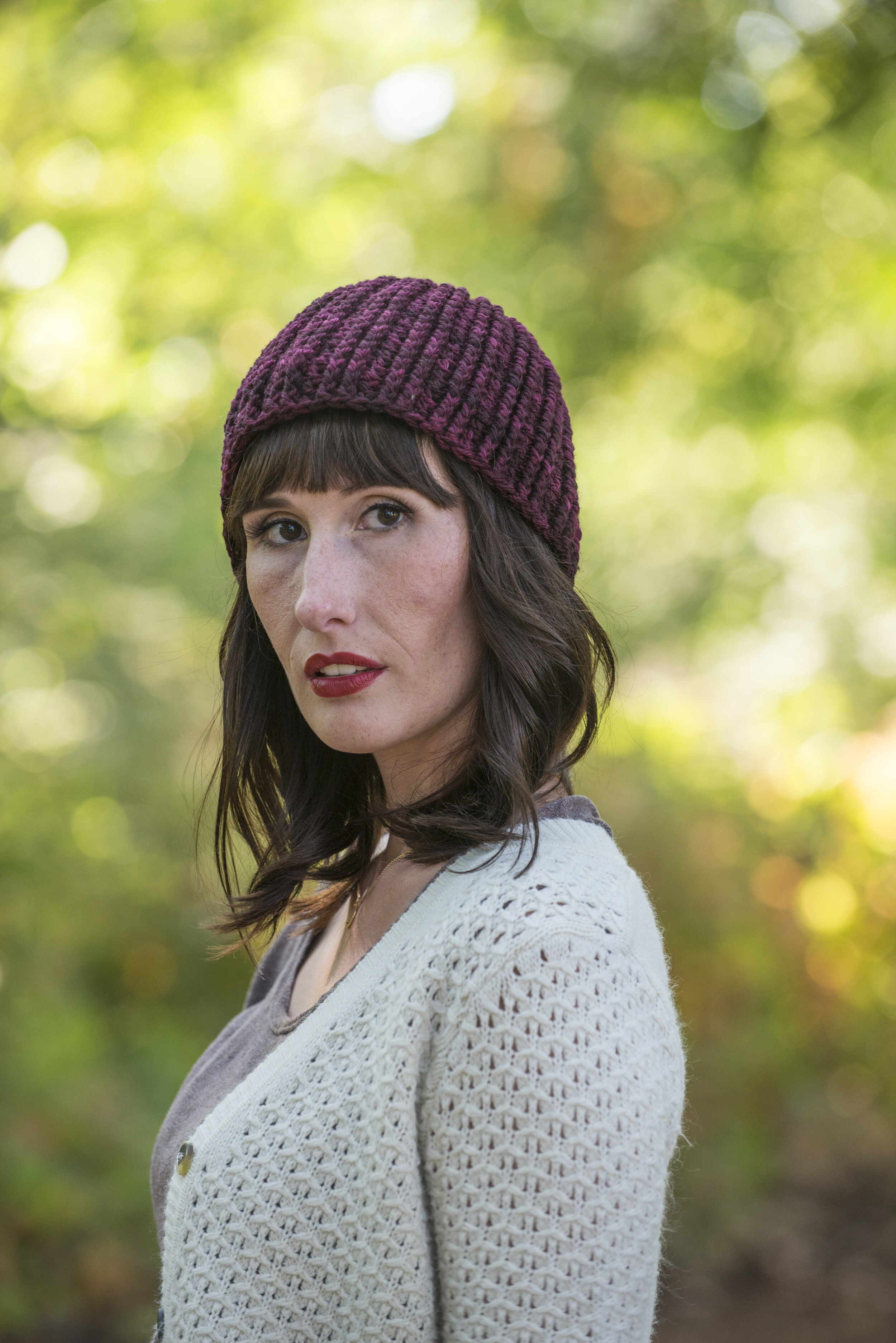  A woman in a white cardigan wearing a deep, wine-colored beanie looking over her shoulder. 