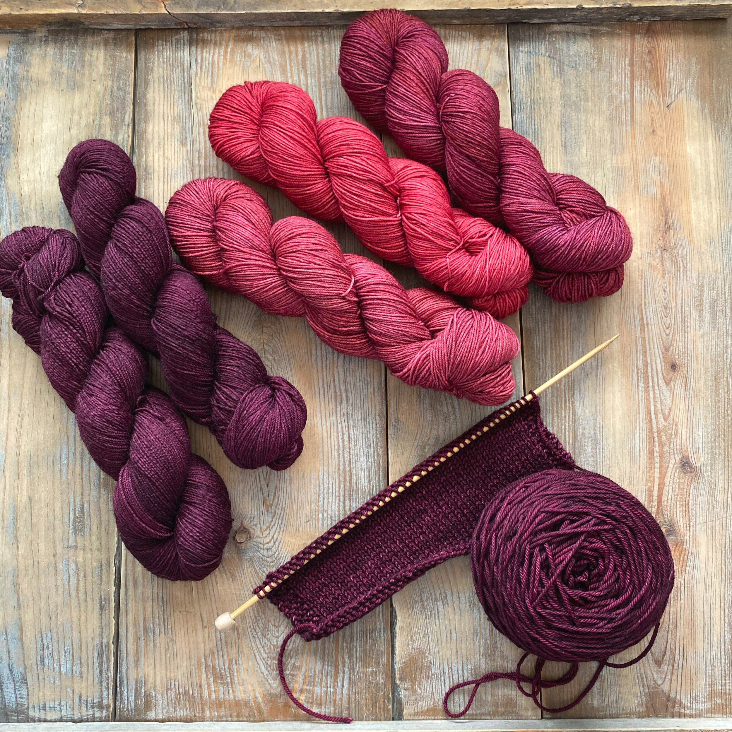 Five skeins of pink and maroon yarn over a cake of maroon yarn on knitting needles.