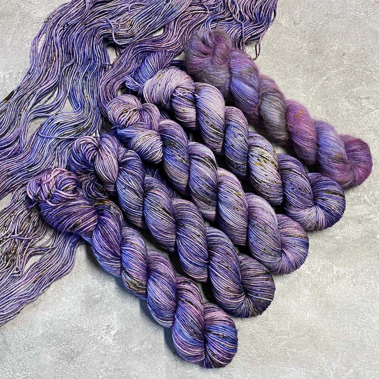 Four skeins of purple yarn in various weights with lighter purple, gray, and yellow speckles sitting on top of spread out yarn in the same color, dyed by Totally Rad Yarn.