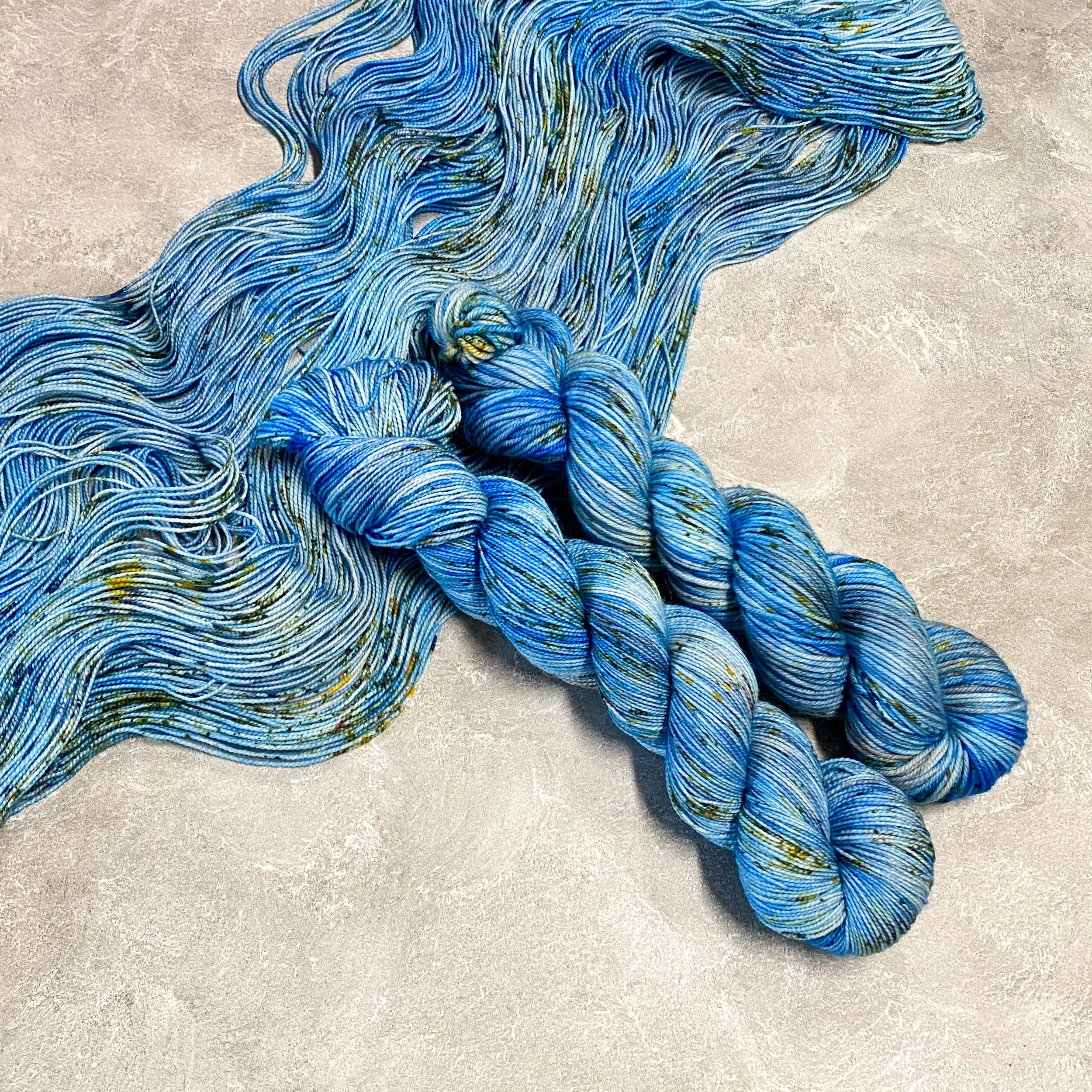 Two skeins of light blue yarn with gray and yellow speckles sitting on top of spread out yarn in the same color, dyed by Totally Rad Yarn.