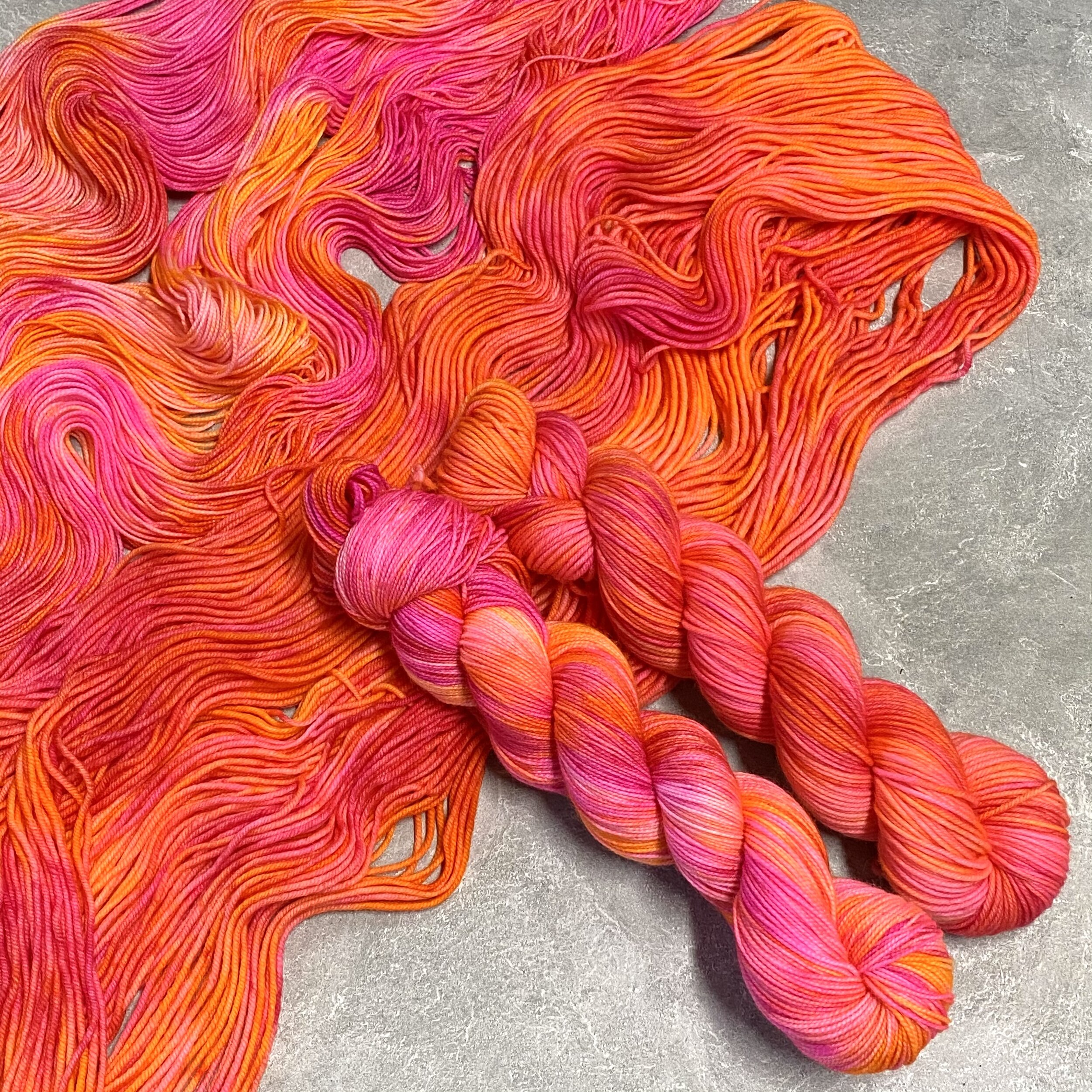 Two skeins of pink and orange tonal yarn sitting on top of spread out yarn in the same color, dyed by Totally Rad Yarn.
