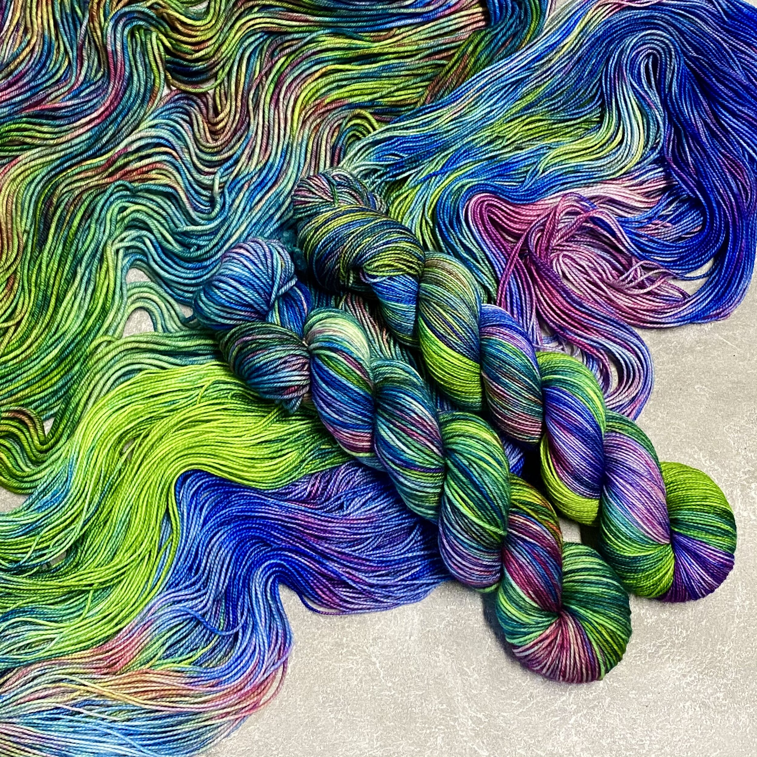 Two skeins of variegated yarn with pink, purple, blue, and green colors sitting on top of spread out yarn in the same color, dyed by Totally Rad Yarn.
