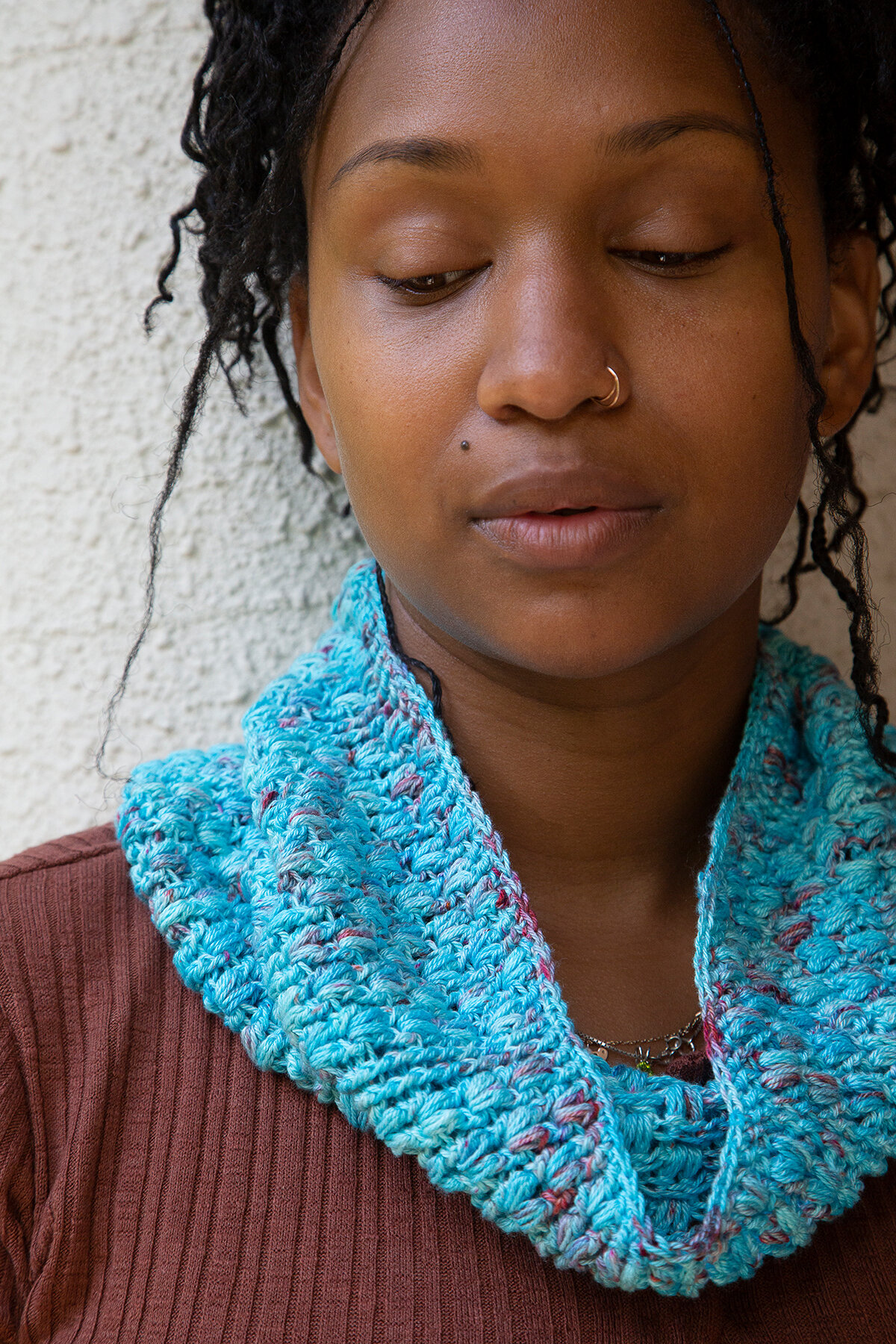 A woman looks downwards while wearing a crocheted Seashell Cowl made of blue yarn around her neck.