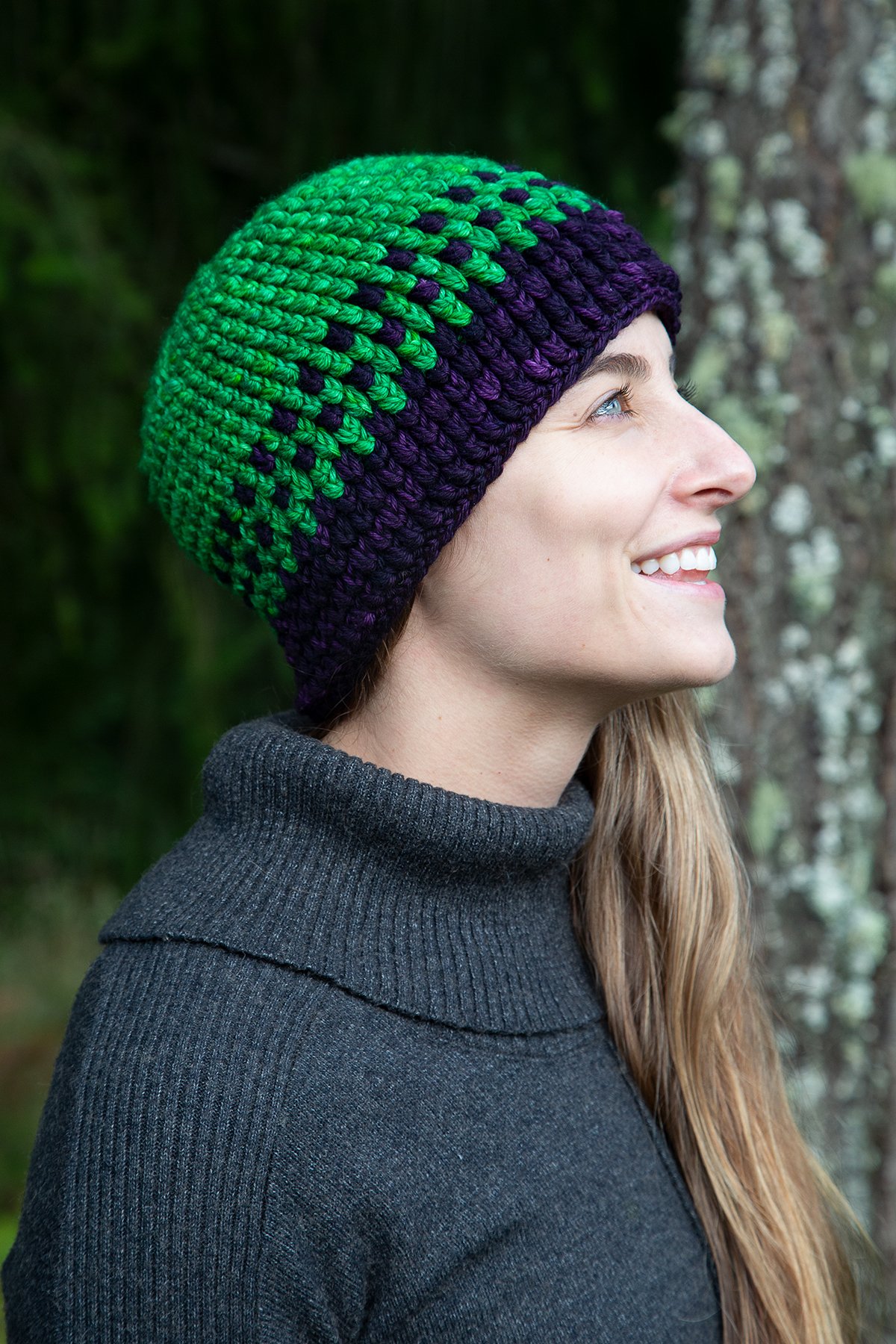 A woman smiles and looks upwards while wearing the green and purple Yarn Dragon Beanie.