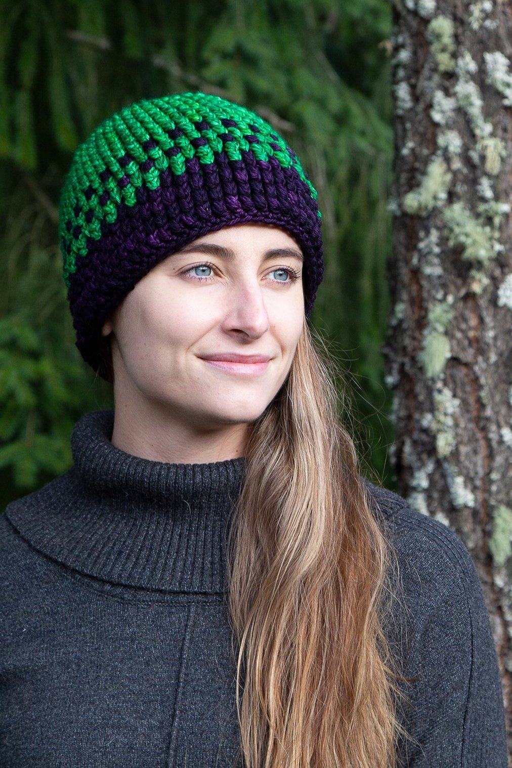 A woman with long brown hair wears the crocheted Yarn Dragon Beanie made out of green and purple yarn.