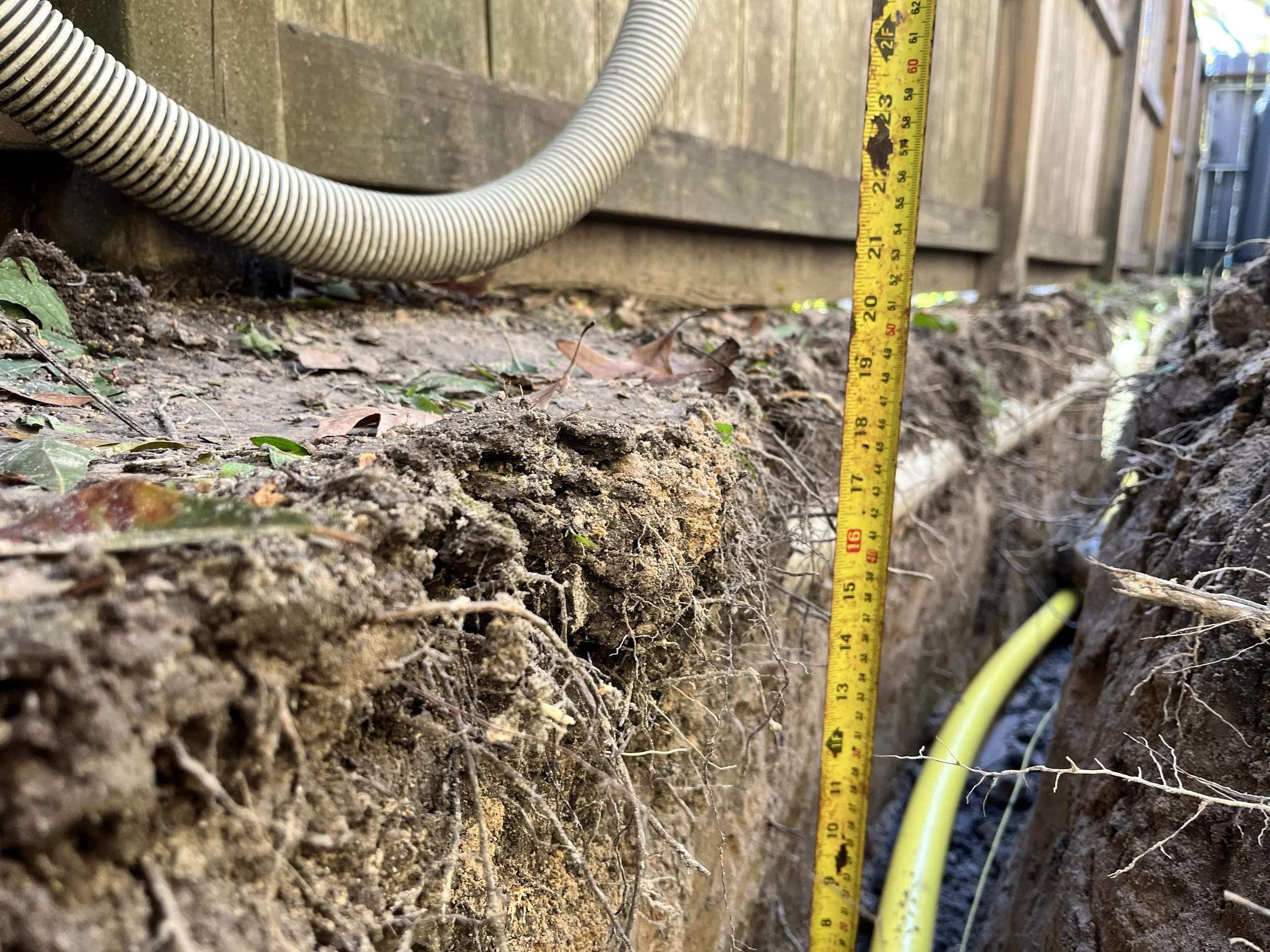 A close-up view of a dirt ground with a yellow measuring tape standing vertically and a white flexible corrugated pipe, both next to a wooden fence. The measuring tape indicates approximately 21 inches.