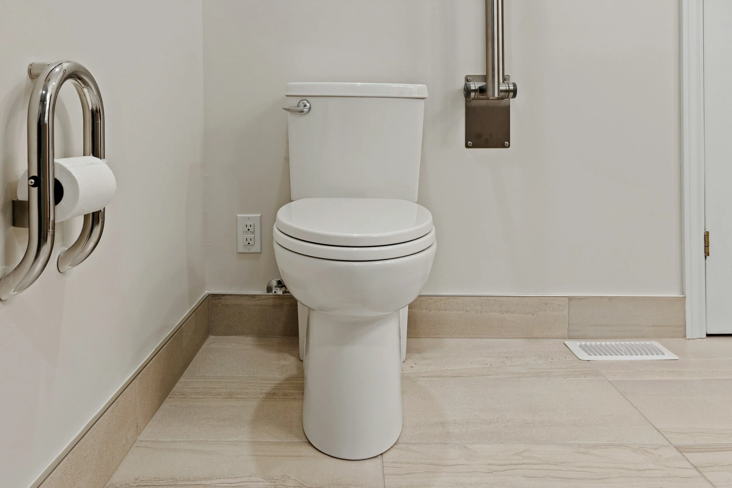 A clean and modern bathroom with a toilet, a fold-up metal grab bar on the left wall, a toilet paper roll on a metal holder, and a floor vent near the baseboard.