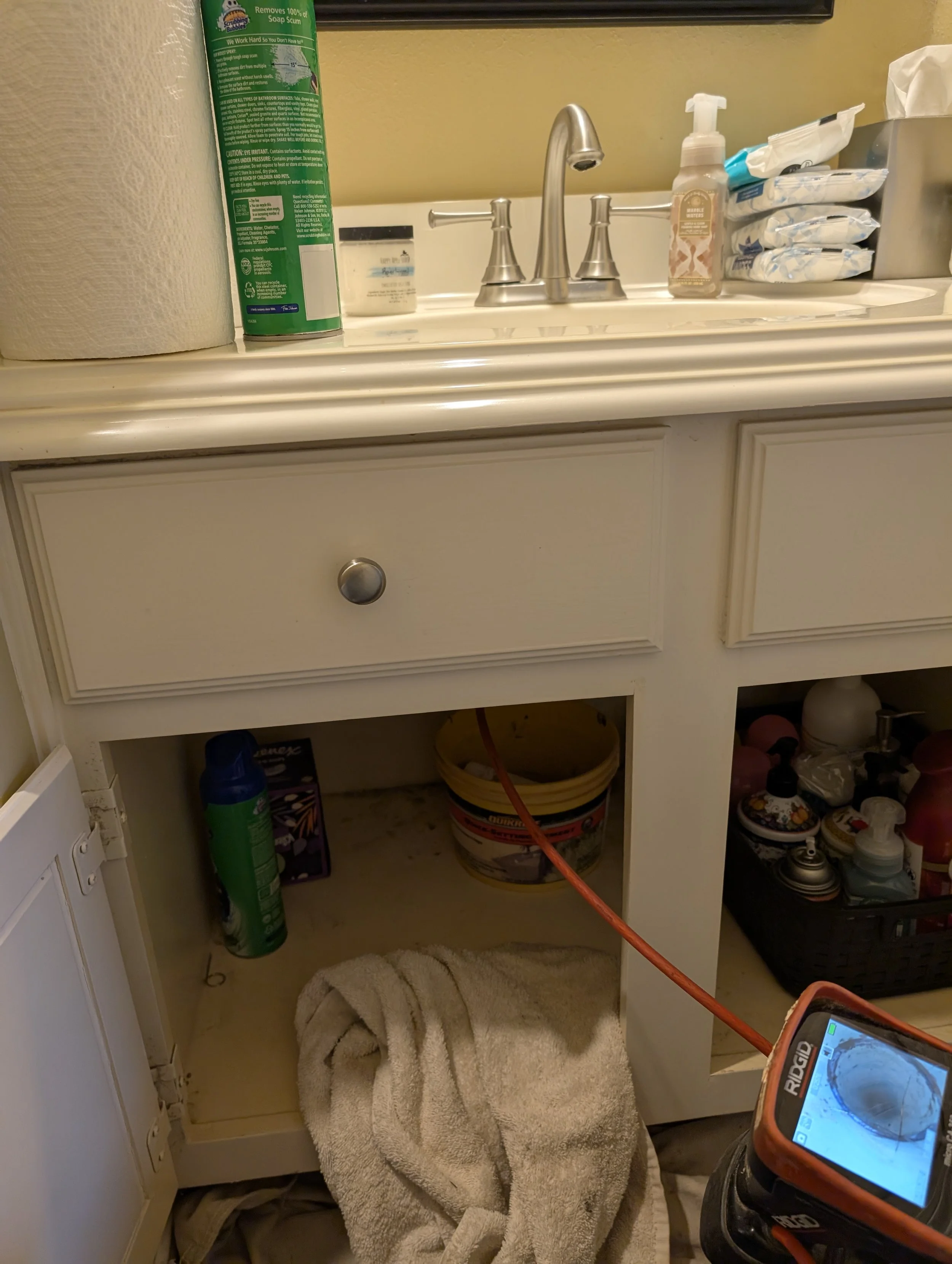 Bathroom vanity with sink, soap dispenser, wipes, and cleaning supplies, with an open cabinet shelf containing a towel, cleaning products, and cleaning equipment, in a bathroom setting.