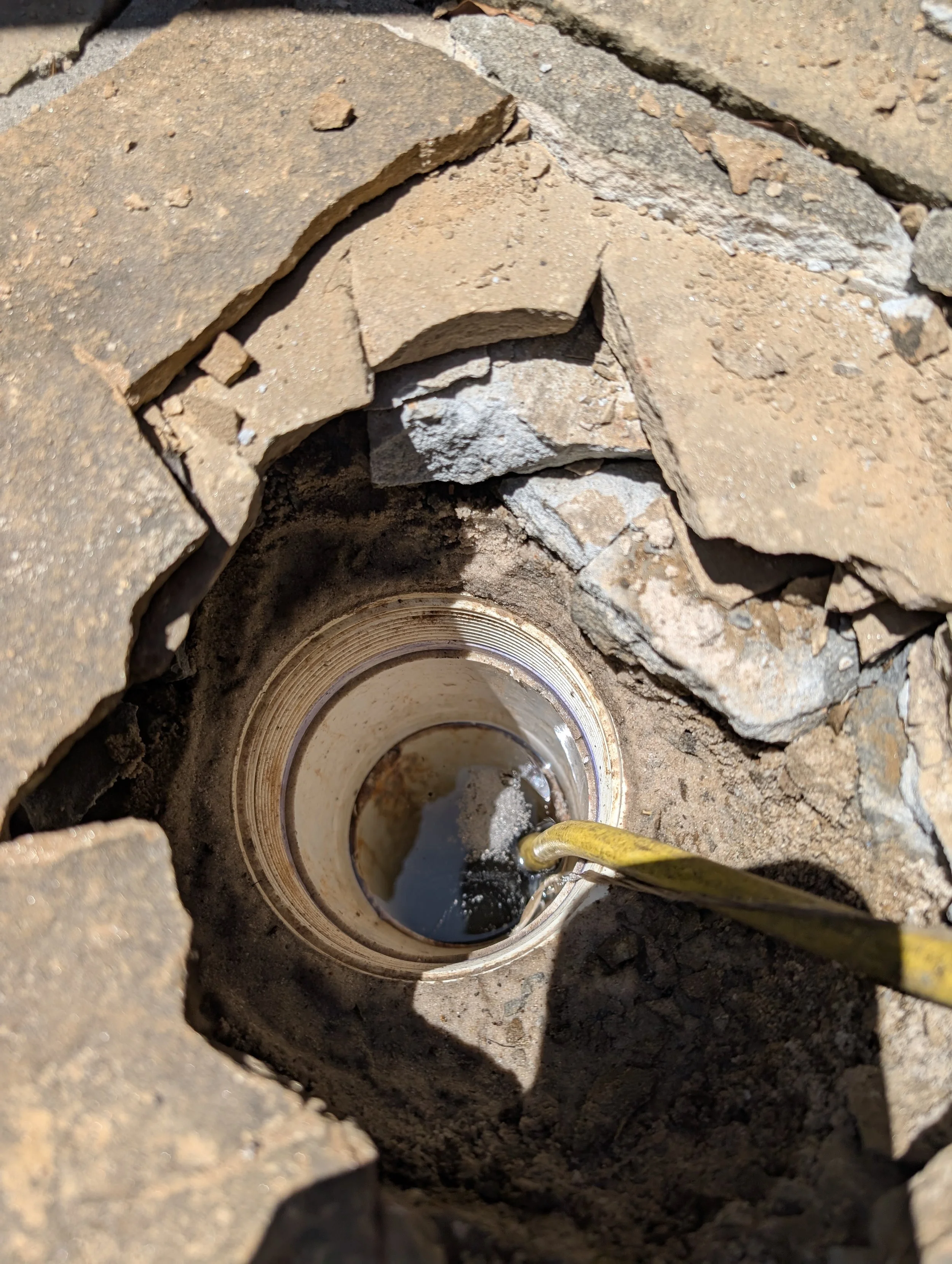 Close-up of a sewer or pipe opening surrounded by bricks and dirt, with a yellow hose or pipe inserted into the opening.
