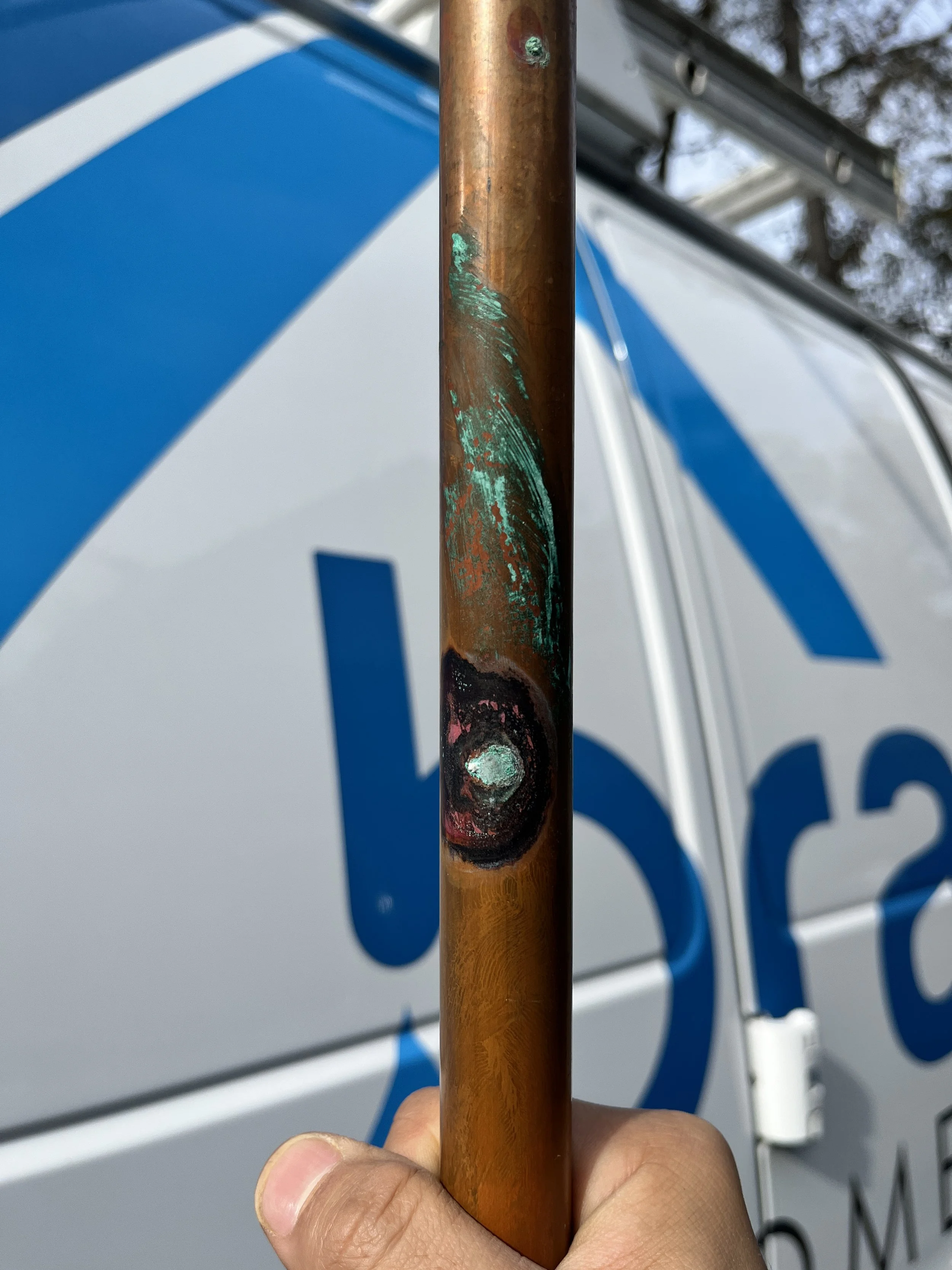 Close-up of a rusty, weathered copper pole with peeling green paint being held in front of a white and blue service vehicle.