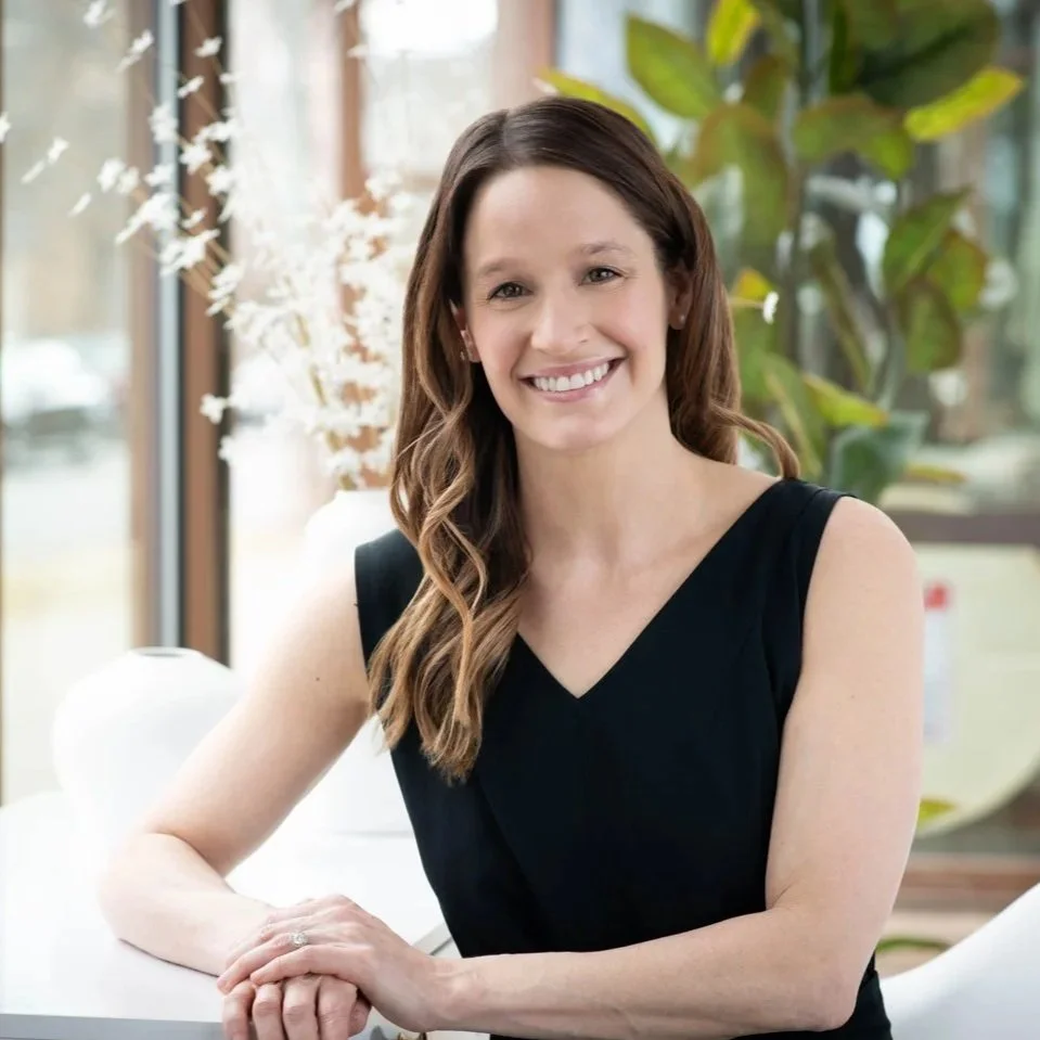 Personal branding portrait of a smiling woman in a black sleeveless top, seated at a table in a bright interior.