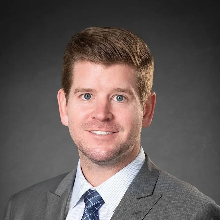 Corporate headshot of a man in a gray suit and patterned tie against a dark studio background.