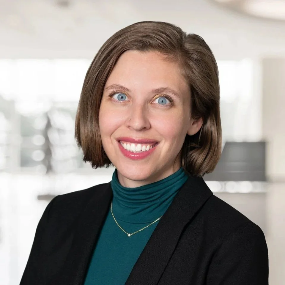 Professional headshot of a smiling woman in a black blazer and teal blouse in a bright indoor setting.
