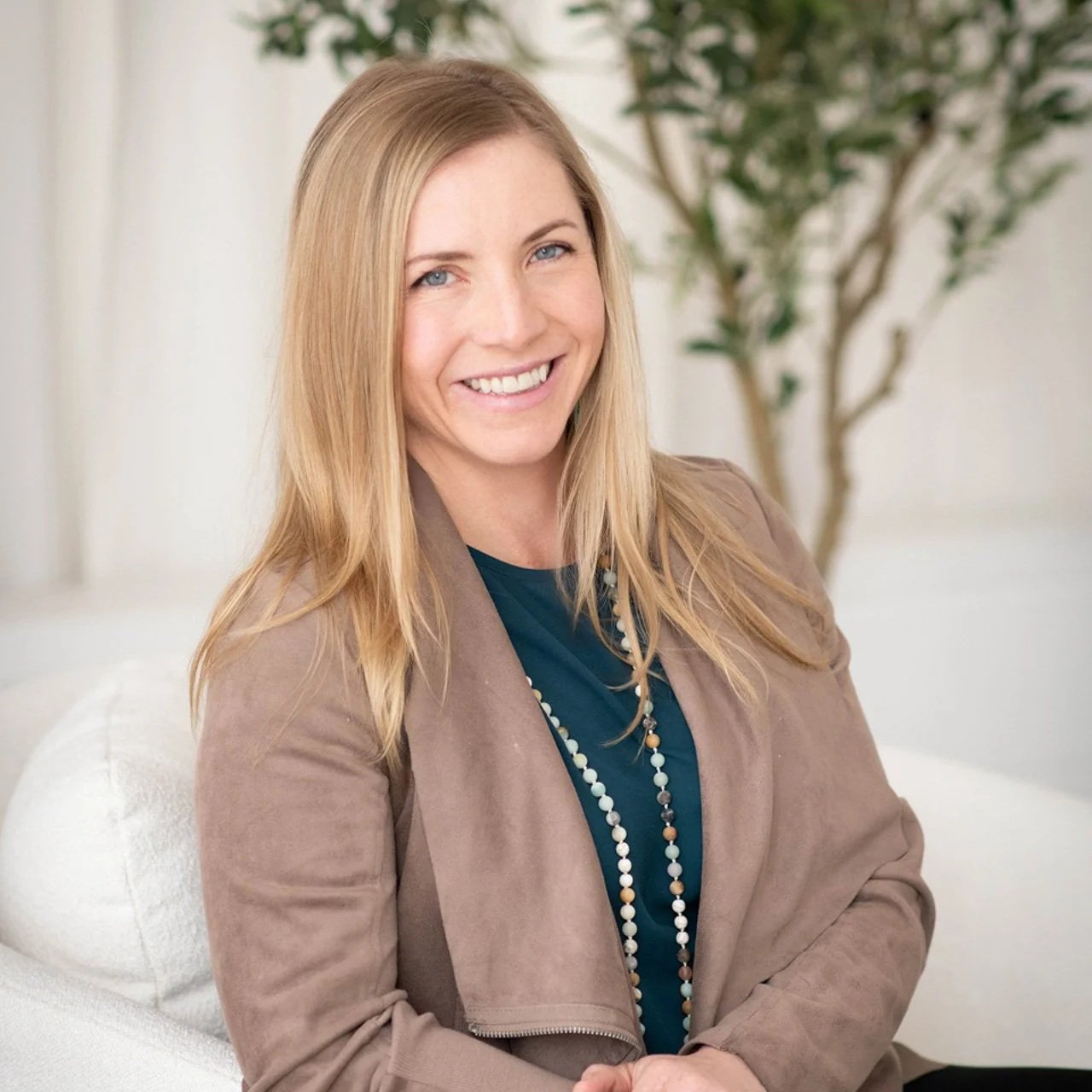 Personal branding portrait of a smiling woman wearing a tan blazer over a teal blouse, seated beside greenery.