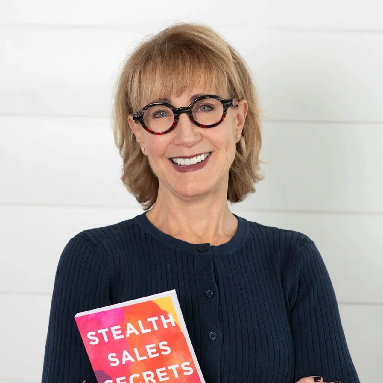Personal branding portrait of a smiling woman wearing glasses and holding “Stealth Sales Secrets” against a light studio background.