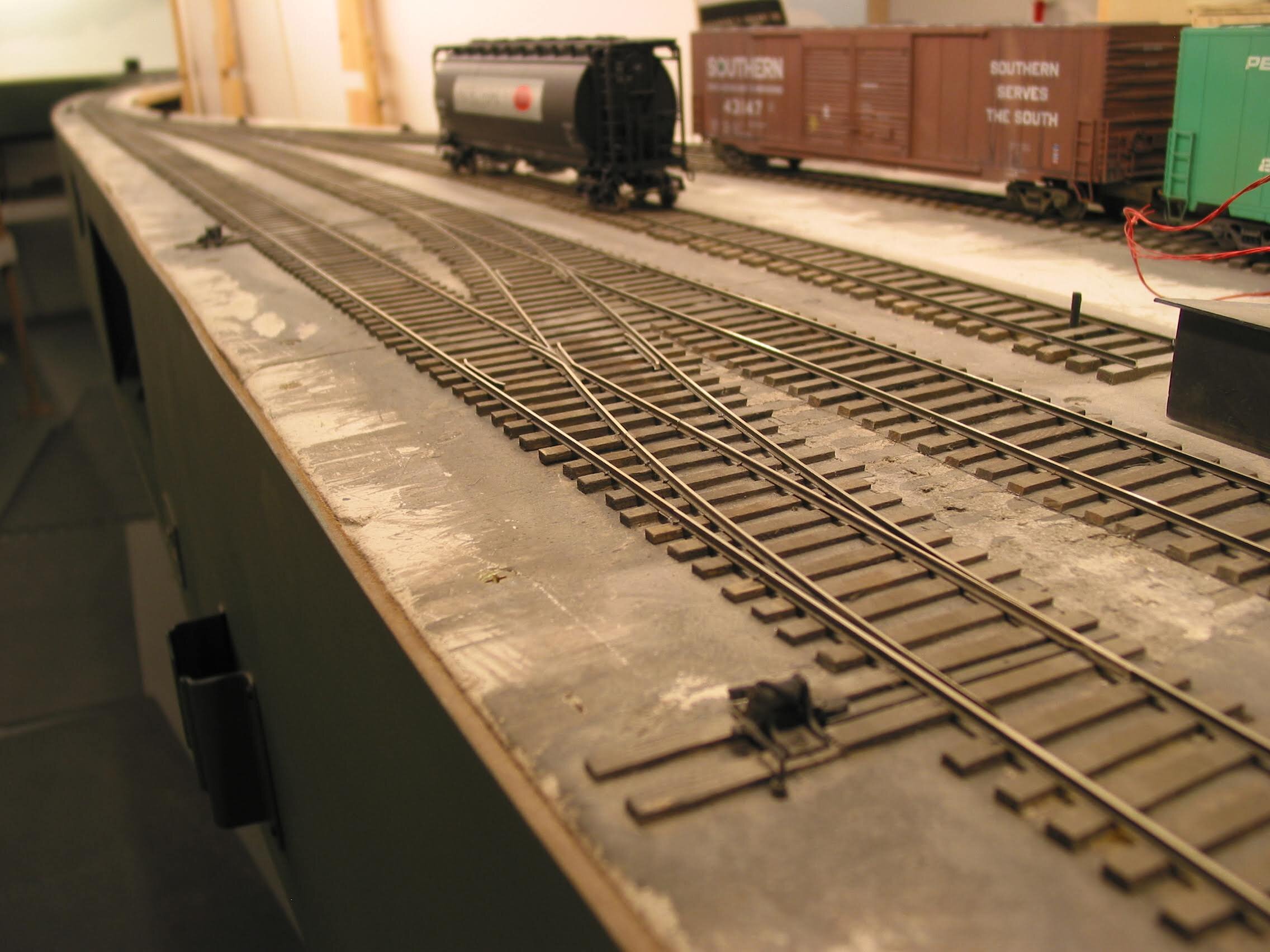  In Fillmore Heights Industrial the trackage is shallow, and high (intentionally). I chose to use HO scale Caboose ground throws with a crank mechanism to an underside two-leaf micro switch. Throwing the turnout changes frog polarity and also lights 