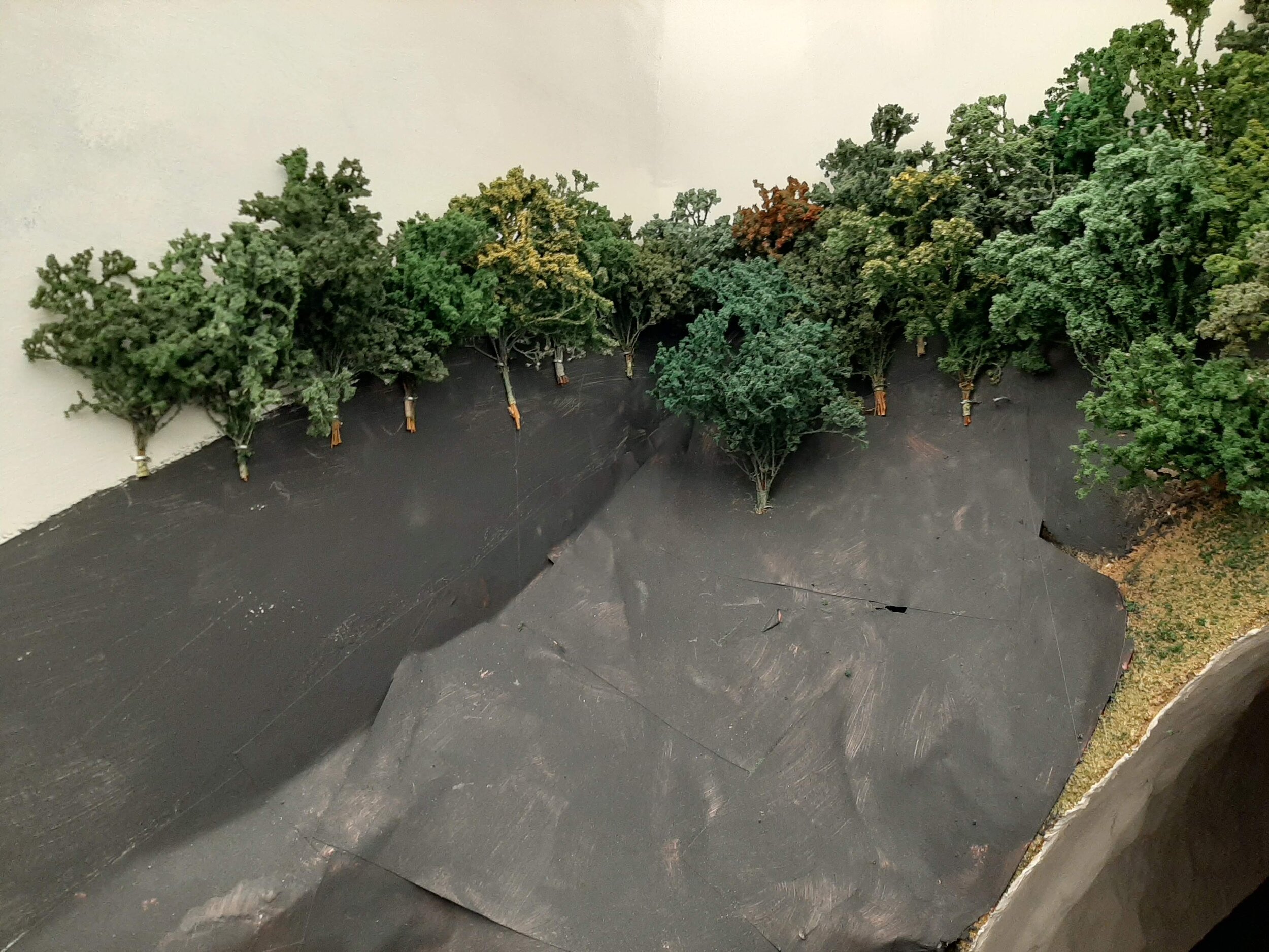  Trees are being “panted” by stapling them to the backdrop/wall. The black paint extends well above the actual landform, thus expanding the scene. And planting smaller trees at the ridge line and increasing in tree size as I progress down the hillsid