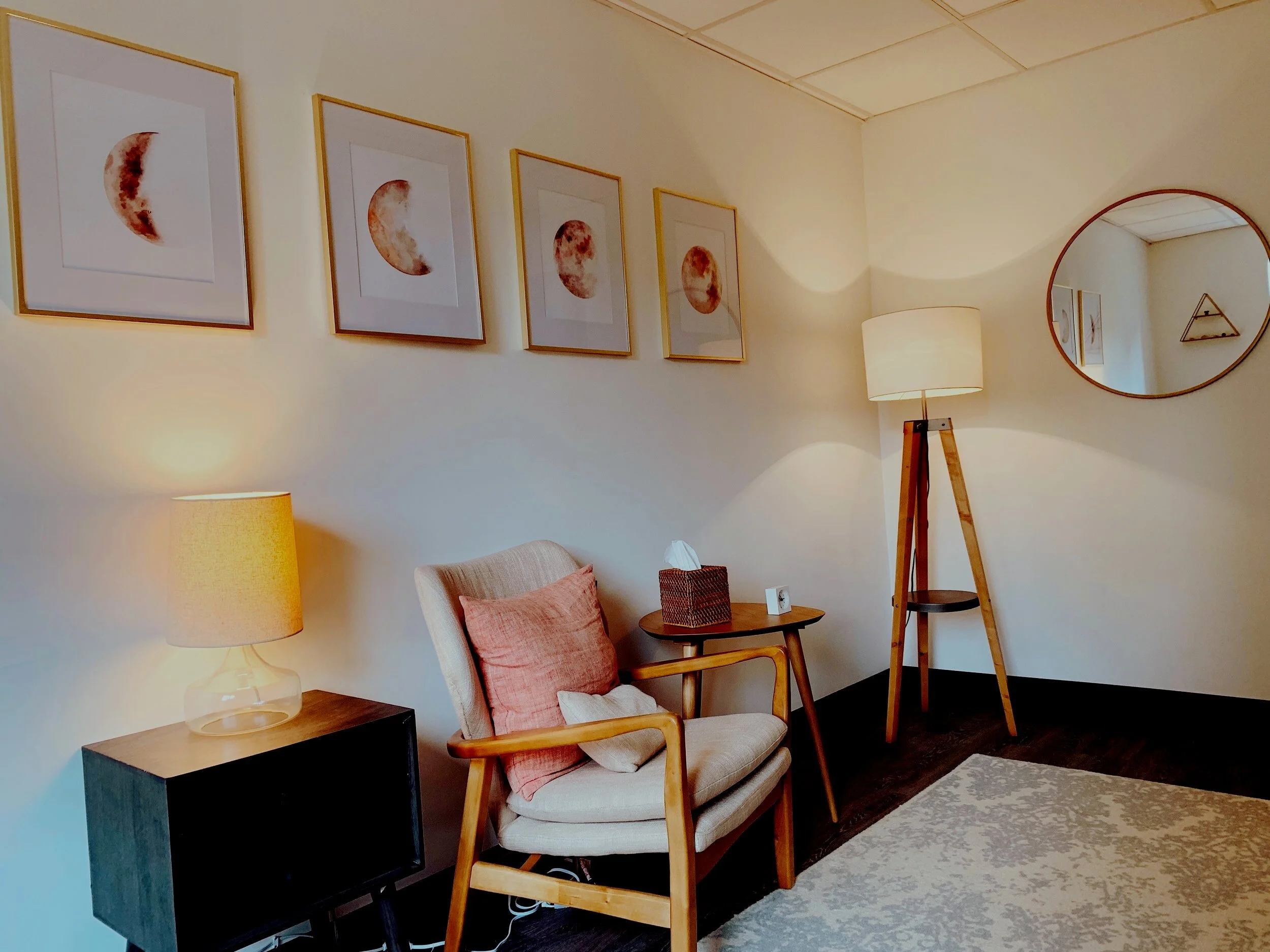 Living room with framed moon phase artwork, a beige armchair with pink pillows, a wooden side table with tissues and a clock, a standing floor lamp, a small black TV on a wooden stand, a mirror on the wall, and part of a cream-colored rug.