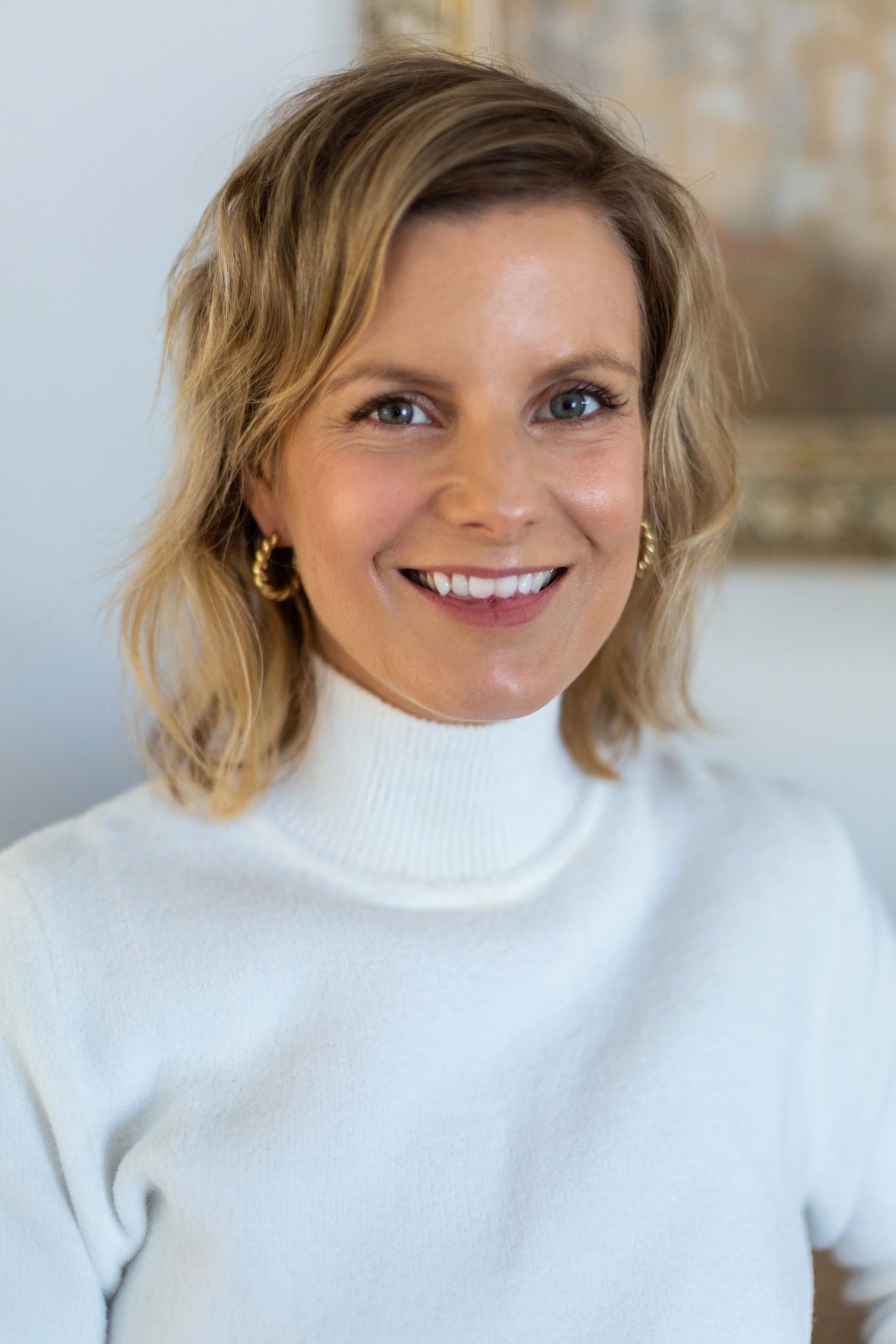 A smiling woman with shoulder-length blonde hair, wearing a white turtleneck sweater and gold hoop earrings, standing indoors with a blurred background.