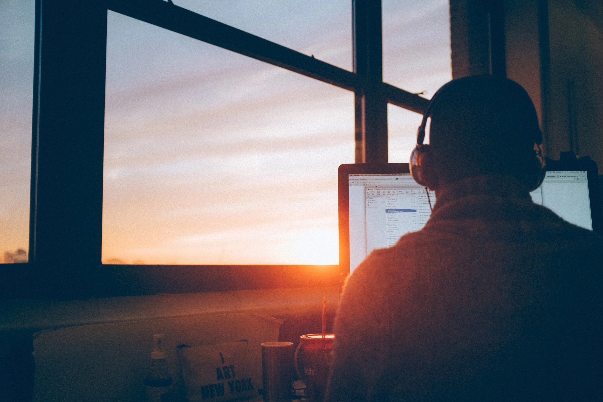 Person with headphones working on a computer at a desk near large windows during sunset.