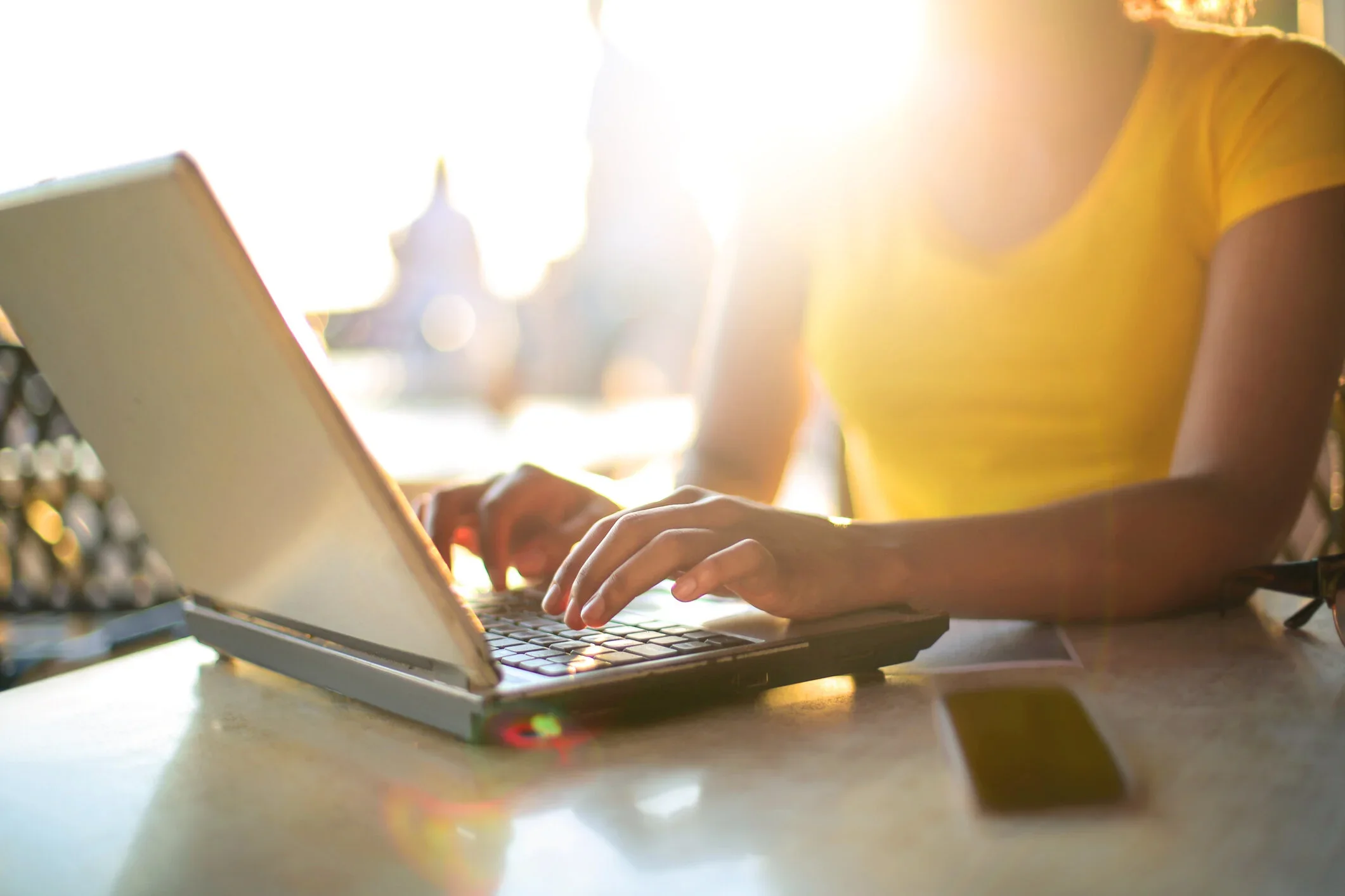 Person wearing a yellow shirt working on a laptop at a table in bright sunlight.