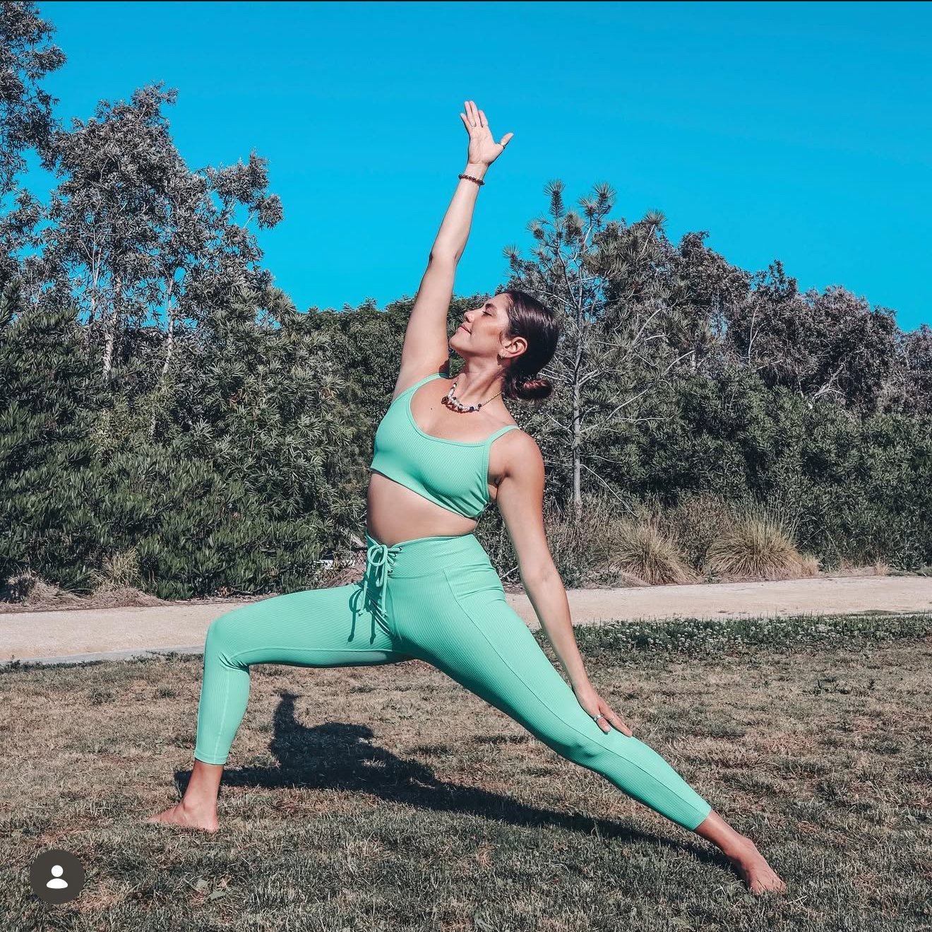 A woman practicing yoga outdoors on grass, wearing a mint green athletic outfit, with trees and blue sky in the background.