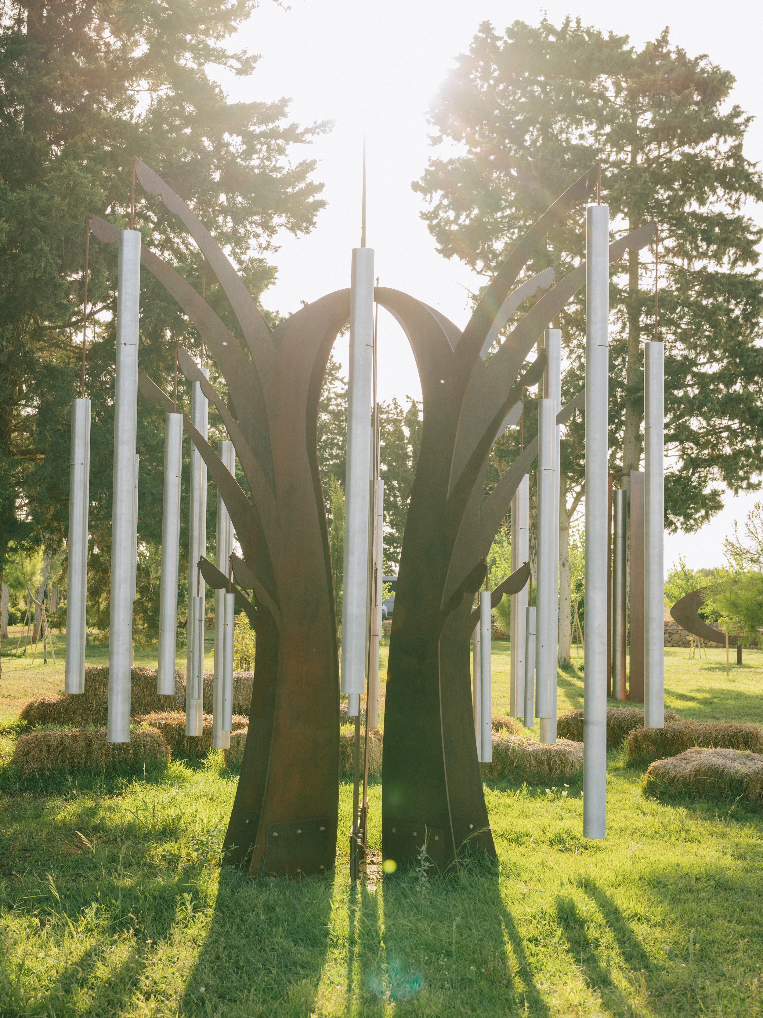 Outdoor sculpture resembling two large stylized trees with black trunks and branches, surrounded by metal tubes hanging vertically and hay bales, with trees and sunlight in the background.